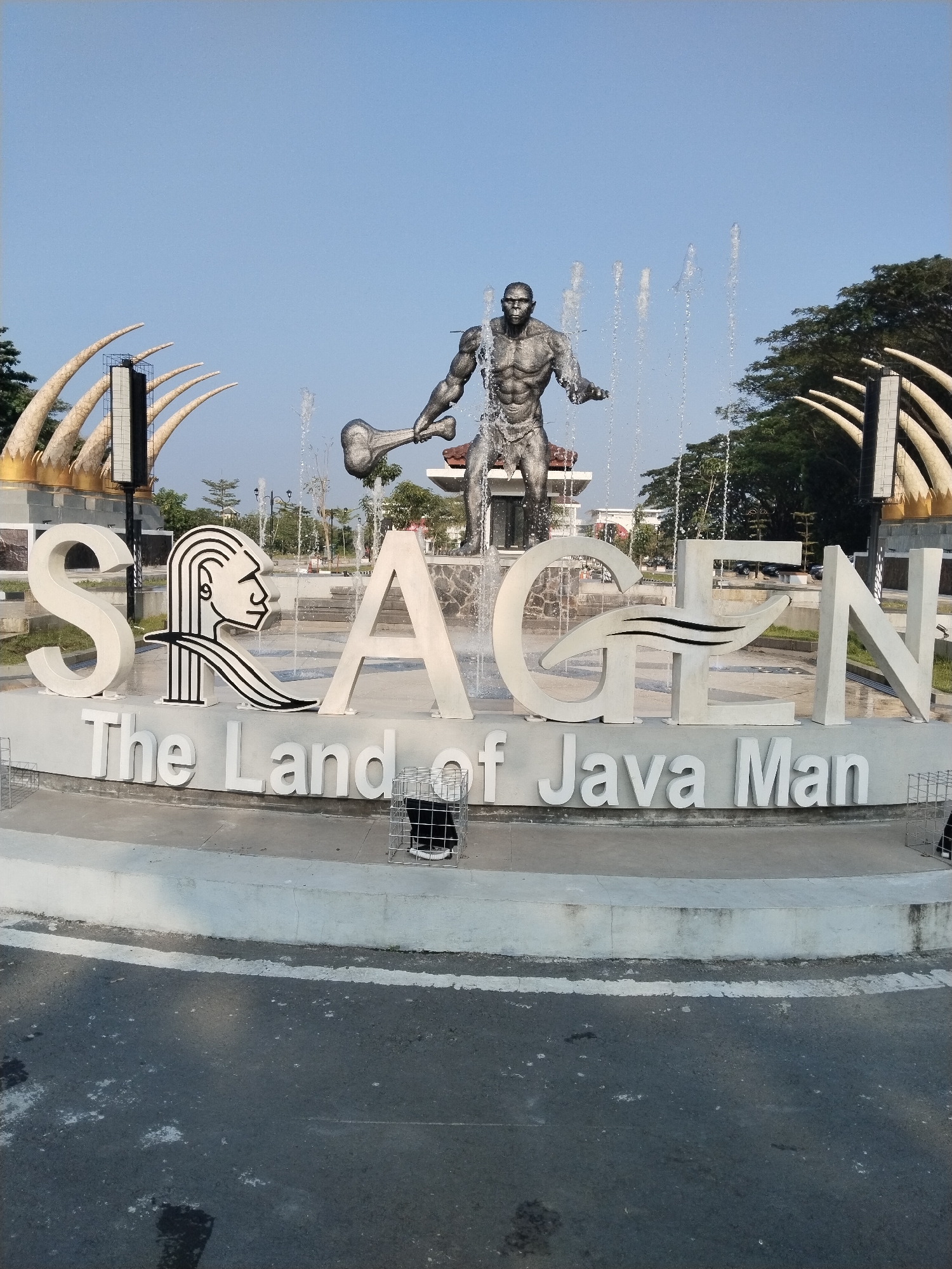 A wide shot captures a striking monument in Sragen Kulon, Indonesia, bathed in clear, bright daylight under a cloudless blue sky. The central feature is a large, multi-component sign spelling "SRAGEN," with "The Land of Java Man" written beneath it in smaller letters.
The letters of "SRAGEN" are prominent and white. The 'R' is uniquely stylized as a black outline of a human profile, possibly representing Java Man, facing left. The 'G' incorporates a black, wavy design within its structure. Behind and around the monument, several tall water fountains spray upwards, creating a dynamic backdrop for a large, dark-colored statue of a muscular, primitive-looking man. This figure, likely representing Java Man, stands firmly, holding a large club or bone in his right hand.
In the background, on either side of the monument, are large, bone-like or tusk-like structures, light in color and curving upwards, reinforcing the prehistoric theme. Green trees are visible beyond these structures, and in the far distance, other buildings and elements of the urban landscape can be discerned.
The foreground shows a paved road surface with some wear and white markings. To the right of the monument, a black cat is visible, confined within a small, light-colored wire cage. Another similar empty cage is present on the left side. The scene effectively conveys the historical and archaeological significance of Sragen as "The Land of Java Man."