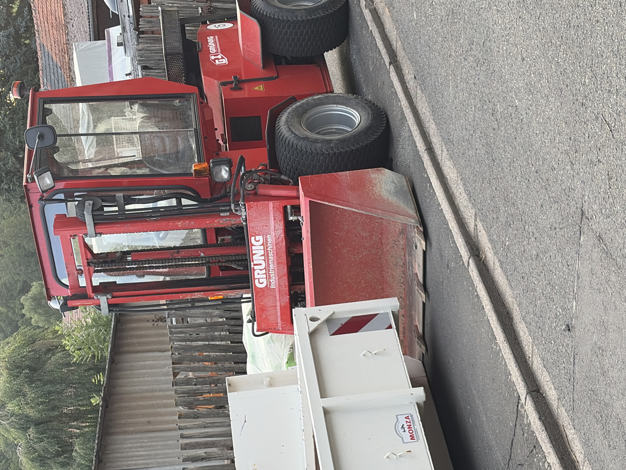 A red GRÜNIG industrial forklift is parked on a paved road next to a white trailer. The forklift has a cab with large windows, a red lift mechanism, and a bucket attachment. The trailer is white and appears to be carrying some materials covered in green plastic. The scene is outdoors, with trees and buildings visible in the background. The road has a curb and some cracks in the asphalt. There are no people visible in the image.