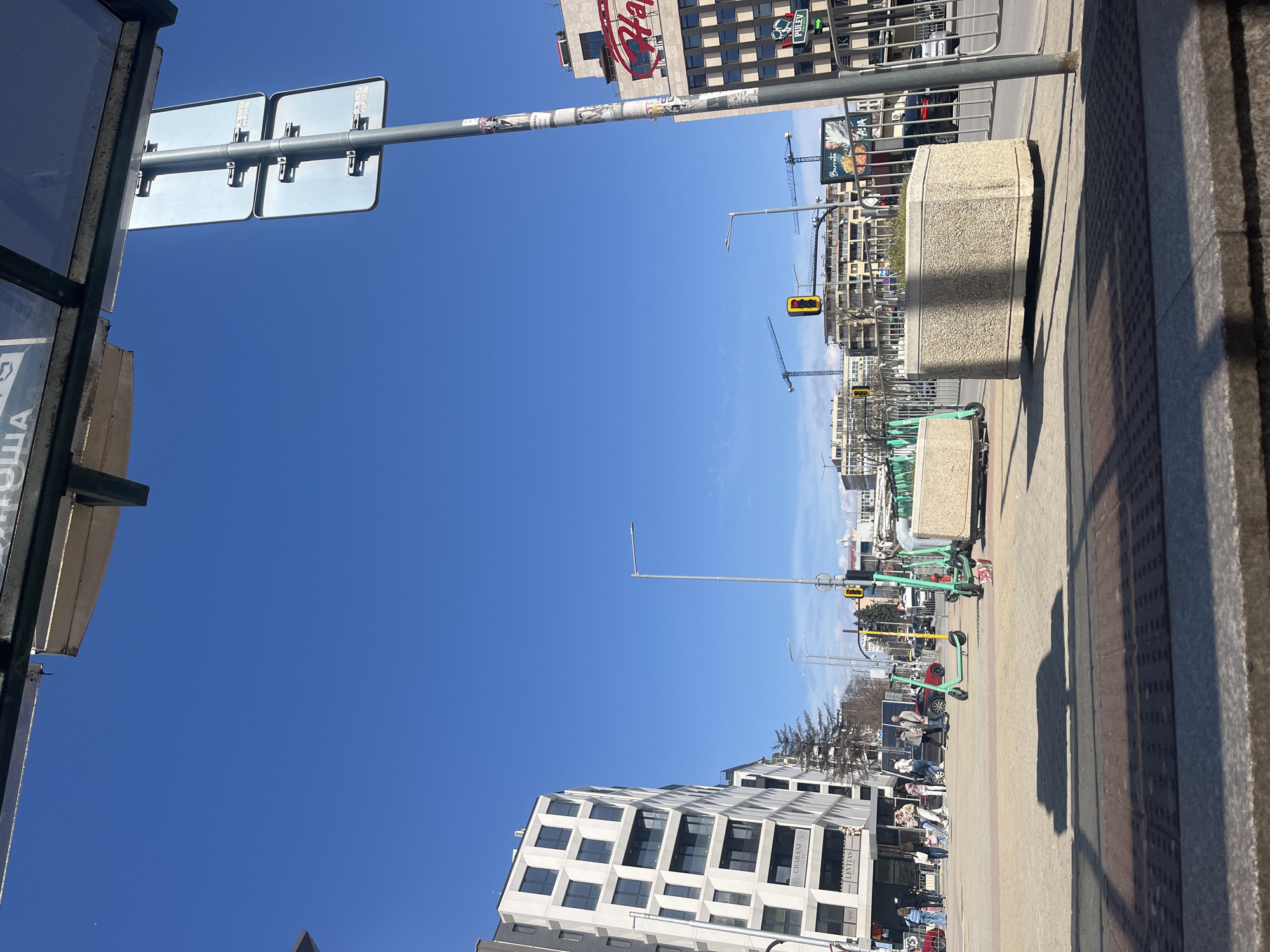 The image depicts a street scene in Sofia, Bulgaria. The camera faces down a pedestrian walkway. On the right, the walkway is bordered by the edge of a building or structure. Several electric scooters, mostly green, are parked along the walkway. Further down, pedestrians are visible sitting at tables outside a building with businesses such as "Levitas" and "Gharani". A construction crane is visible in the distance. Above the building, traffic lights are fixed on poles. A building with a large "Har" sign is visible. The sky is clear and blue. Traffic and pedestrians are visible. A street sign is at the top left.