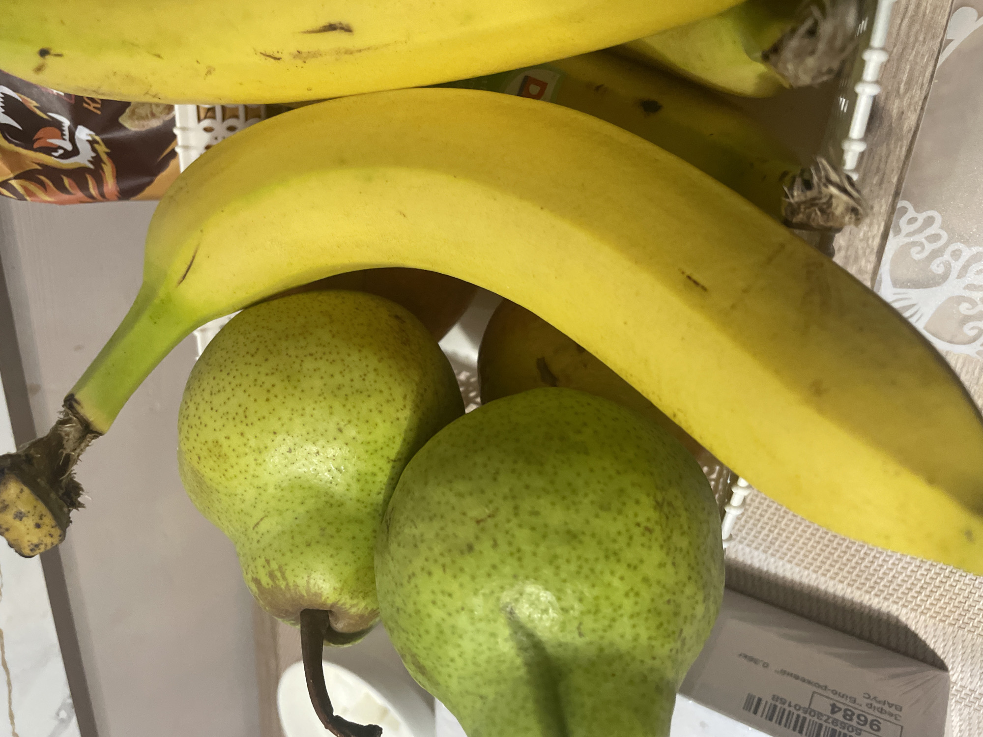The image shows a close-up view of fruit, including pears and bananas, arranged in a white basket. The pears are green and speckled, while the bananas are yellow and appear ripe. Some of the fruit is in focus, while other parts are blurred. The background is out of focus but appears to be an indoor setting with a patterned surface below the basket. There is also a white cardboard box with some text and a barcode visible on the left side of the image. The overall lighting suggests it might be daytime. There are no people or animals visible in the image, nor are there any specific landmarks that would indicate the city of Brovary, Ukraine.