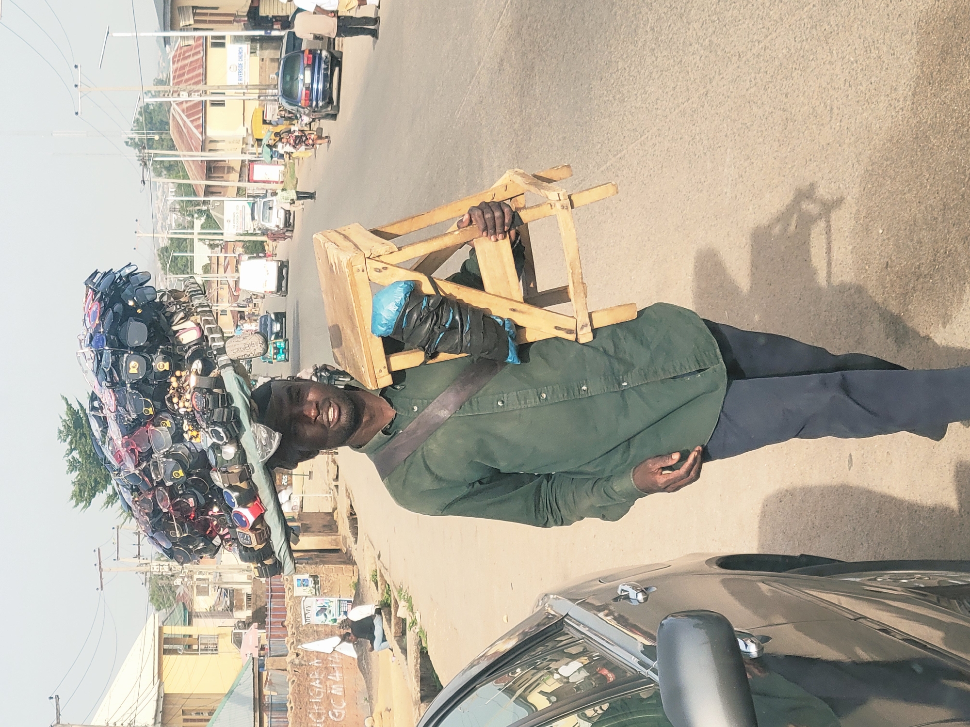 A man stands on a street in Jos, Nigeria, with a wooden chair held over his left shoulder and a display of sunglasses and watches balanced on his head. He wears a dark green button-down shirt, dark pants, and a cap. A brown strap runs across his chest. The street scene includes cars, buildings, and other pedestrians. Graffiti is visible on a wall in the background. The weather appears sunny and bright.