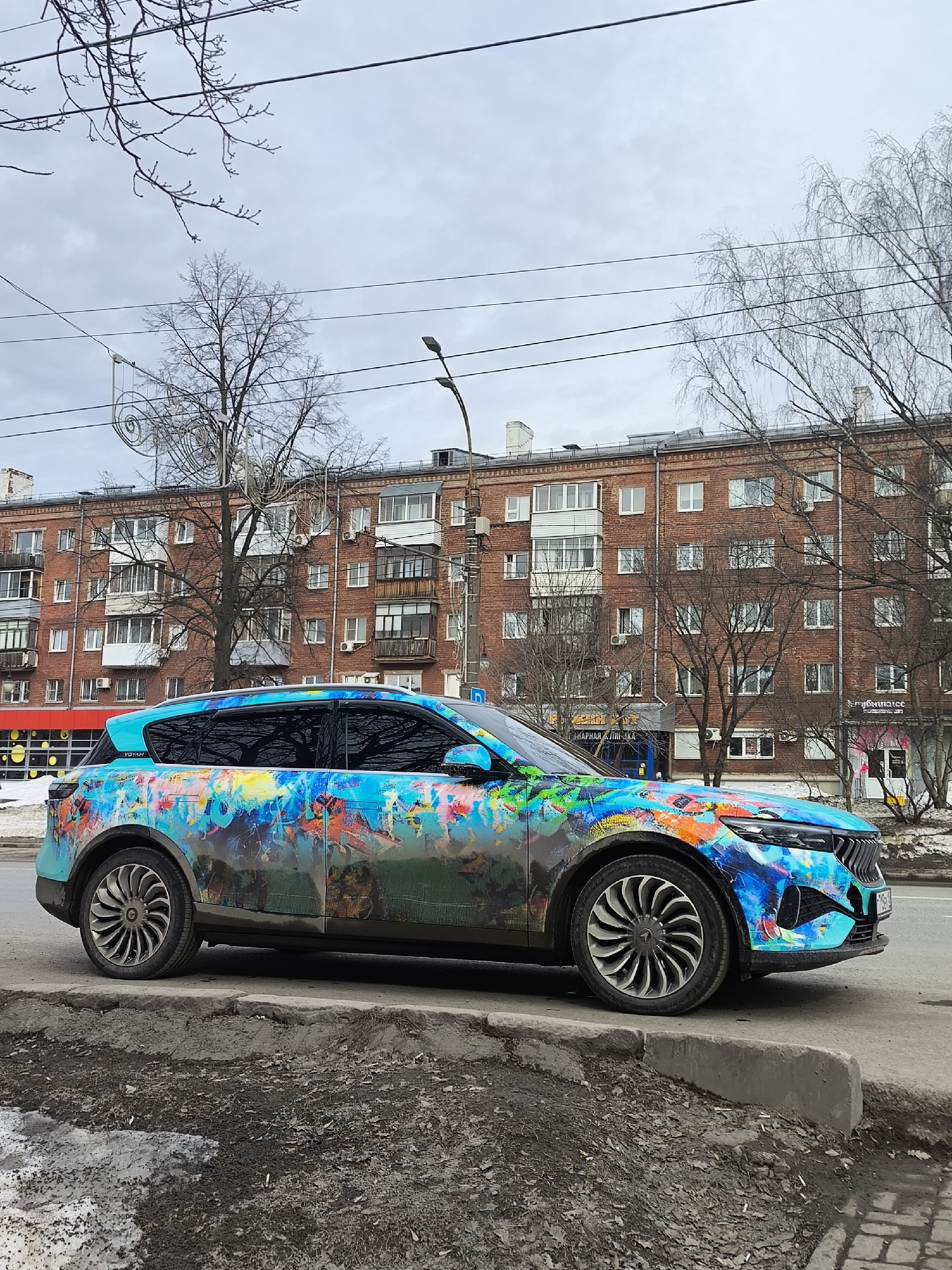 The image shows a Voyah Free car with a colorful, abstract paint job parked on a curb in Izhevsk, Russia. The car is positioned in front of a multi-story brick building with numerous windows and balconies, likely an apartment complex. The season appears to be late winter or early spring, as evidenced by the bare trees and patches of melting snow on the ground. A street light and some commercial buildings are visible in the background. No people are visible in the frame.