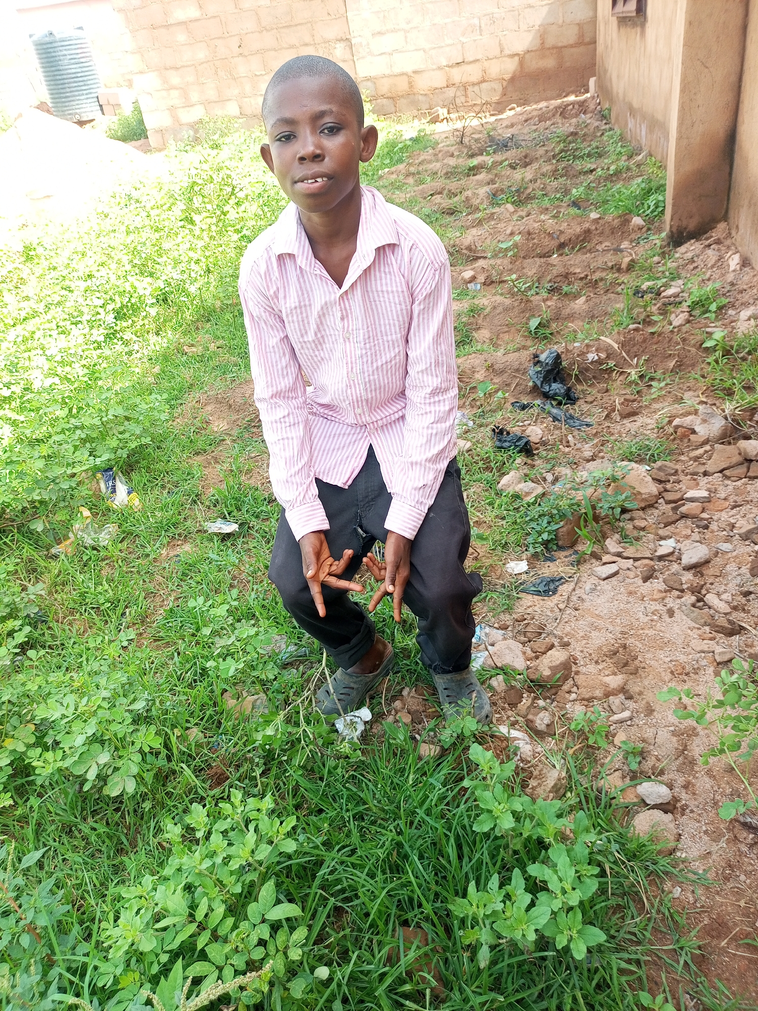 A young boy with dark skin and short hair is sitting on the ground outdoors, with his knees bent and his feet flat on the ground. He is wearing a pink and white striped button-down shirt and black pants. His hands are clasped between his knees. The background consists of a dirt patch with some rocks and sparse vegetation, and a brick wall in the distance. To the left of the boy, there is a patch of green grass with some scattered trash. The scene appears to be in an urban or semi-urban area in Tempure, Nigeria.