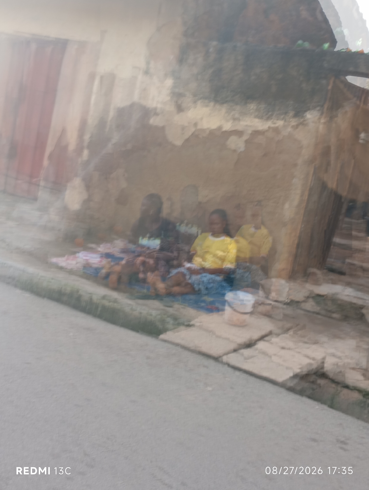 The image shows a street scene in Shendam, Nigeria. Two individuals, likely women, are seated on the ground in front of a weathered building. One person wears a black shirt, while the other is in a yellow shirt. They appear to be selling goods laid out on cloths on the ground. A white plastic bucket sits near them. The background consists of a textured wall and some debris. The foreground shows a paved street. The overall image is somewhat blurry, suggesting motion or a lack of focus. The timestamp "08/27/2026 17:35" and "REDMI 13C" are visible in the lower corners, indicating the date, time, and device used for capture.