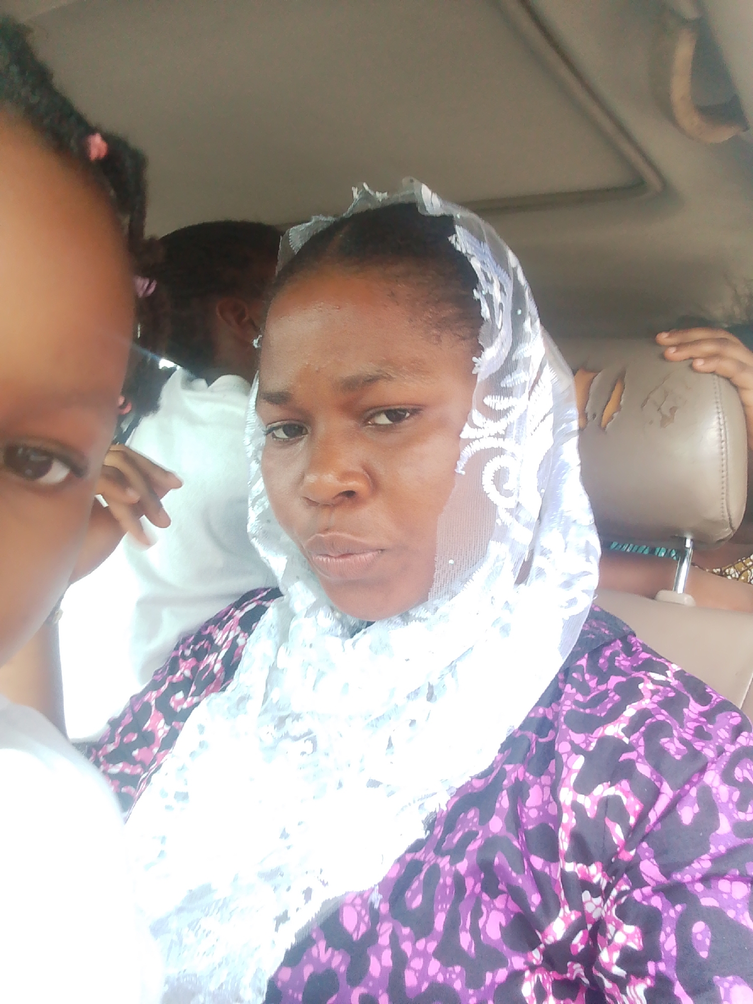 In a car, a woman with dark skin wears a white lace veil over her head and a purple and black patterned dress. Another person, partially visible on the left, has dark braided hair and is out of focus. The car interior features a light-colored headrest with visible damage and a sunroof. The scene is likely in Yenegoa, Nigeria, based on context.