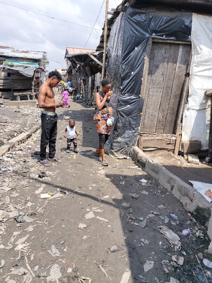 The image depicts a street scene in Oworonshoki, Nigeria, under bright daylight with a partly cloudy sky. The ground consists of an unpaved, uneven dirt path heavily littered with refuse, including plastic fragments, paper, and debris. A low concrete curb runs along the right side of the path, bordering a drainage channel also filled with waste.
In the foreground, three individuals are present. On the left, a shirtless male with dark, curly hair wears dark trousers and light-colored sandals; his light-colored underpants are visible above his waistband. He stands, looking down at an object held in his hands. To his right and slightly forward, a young child, possibly a toddler, stands facing left, wearing a light-colored shirt, patterned shorts, and small shoes. Further to the right and slightly behind the child, a female walks forward (towards the right), wearing a dark sleeveless top, a patterned wrap skirt over dark leggings or trousers, and sandals. She holds a light-colored shoulder bag and has an object held to her mouth.
The environment is characterized by densely packed, rudimentary structures. On the left side of the path, multiple wooden shacks with corrugated metal roofs are visible, some appearing to be constructed from scrap materials, one with a faded light blue section. On the right side, a structure is predominantly constructed from wooden planks and extensively covered with black plastic sheeting (tarpaulin), featuring a rough wooden door. Overhead wires span across the scene. Further down the path, additional indistinct figures and structures are visible, including one person in a pink or purple garment. Shadows indicate the sun is high in the sky, casting shadows towards the right.