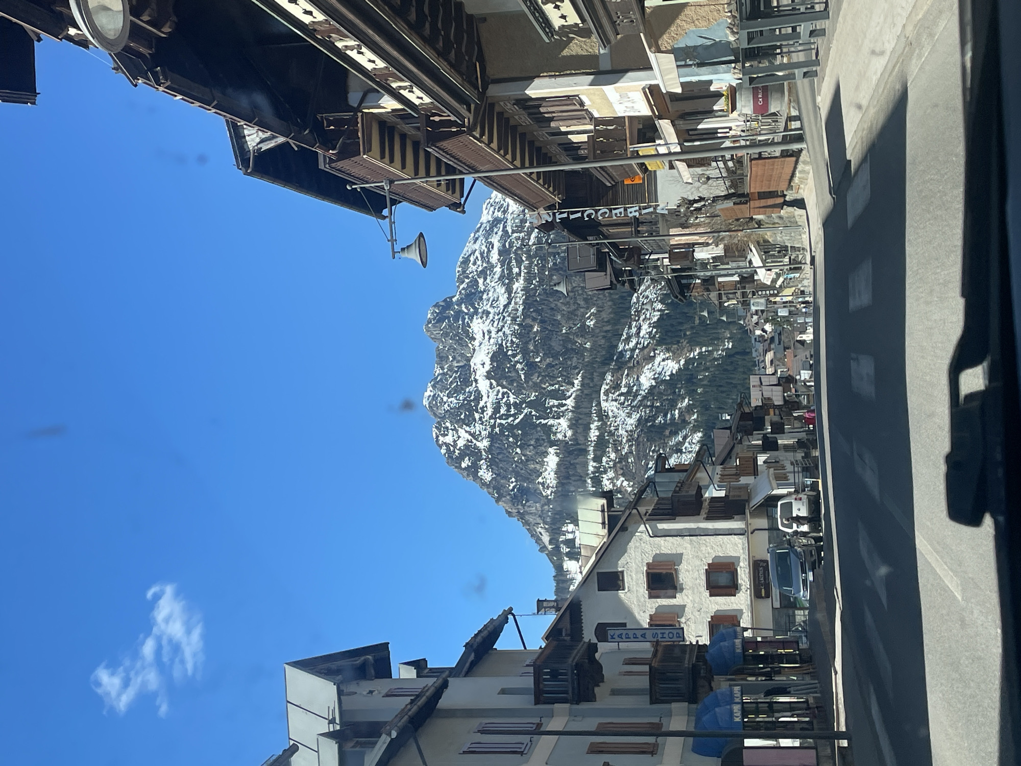 The image shows a view in the town of Sappada, Italy. A snow-covered mountain is visible in the distance, framed by buildings on either side of the street. The buildings appear to be of traditional construction with wooden balconies and window shutters. The street is paved, and there are vehicles parked along the side. The sky is clear and blue with a few clouds. A sign that says "Kappa Shop" is visible on the side of one of the buildings.
