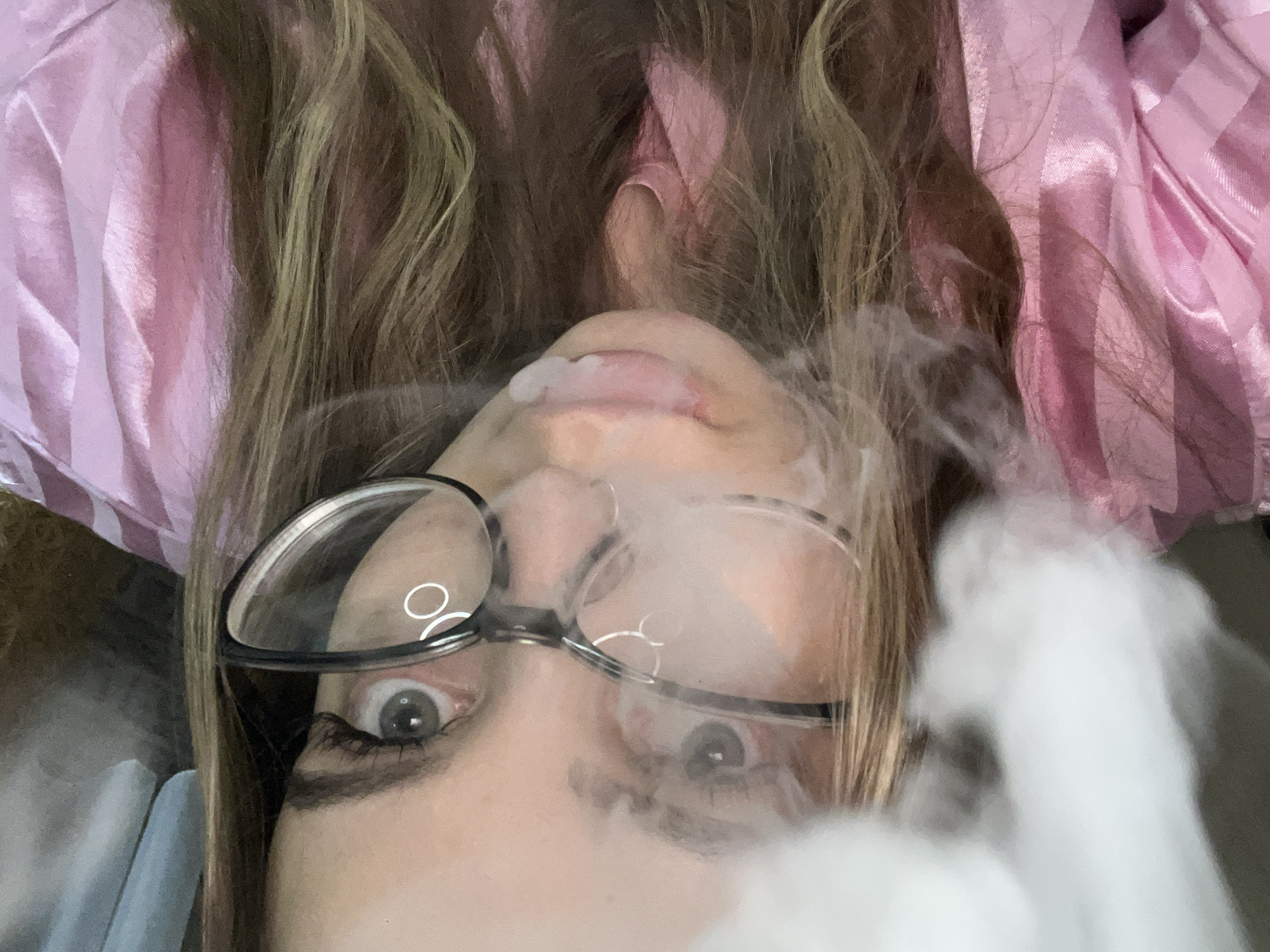 A close-up, tilted image depicts a person wearing glasses exhaling a large plume of white smoke. The individual's face is angled upwards, revealing their nose, mouth, and eyes behind the glasses. The person has fair skin and long, light brown hair that frames their face. A pink, possibly satin or silk, fabric is visible in the background. The location of the image in the city of Novopokrovskaya, Russia is unknown.