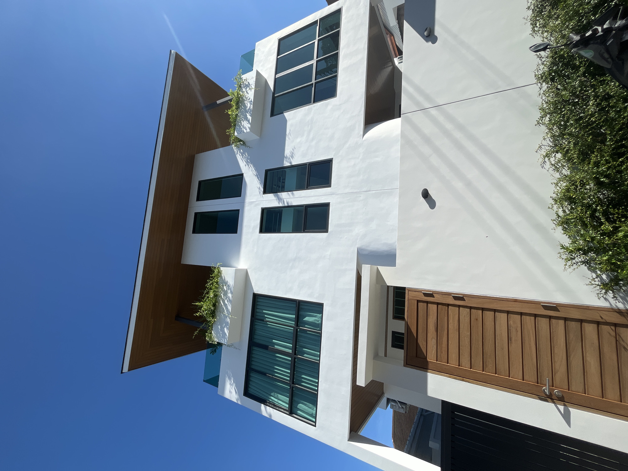 A low-angle, wide shot captures a modern white multi-story house against a bright blue sky in Rawai, Thailand. The house features a distinctive angled wooden-paneled roof with an overhang. Multiple large windows with dark frames are visible on the white facade, some adorned with teal curtains. Planter boxes with greenery sit on the upper levels. A tall, slatted wooden door with a silver handle is prominently placed on the lower section of the house, to the left of the image's center. To the left of the door is a black metal fence. Green bushes line the bottom of the image, and a portion of a black motorcycle with a mirror is visible on the right side. Sunlight casts strong shadows across the white walls of the house.