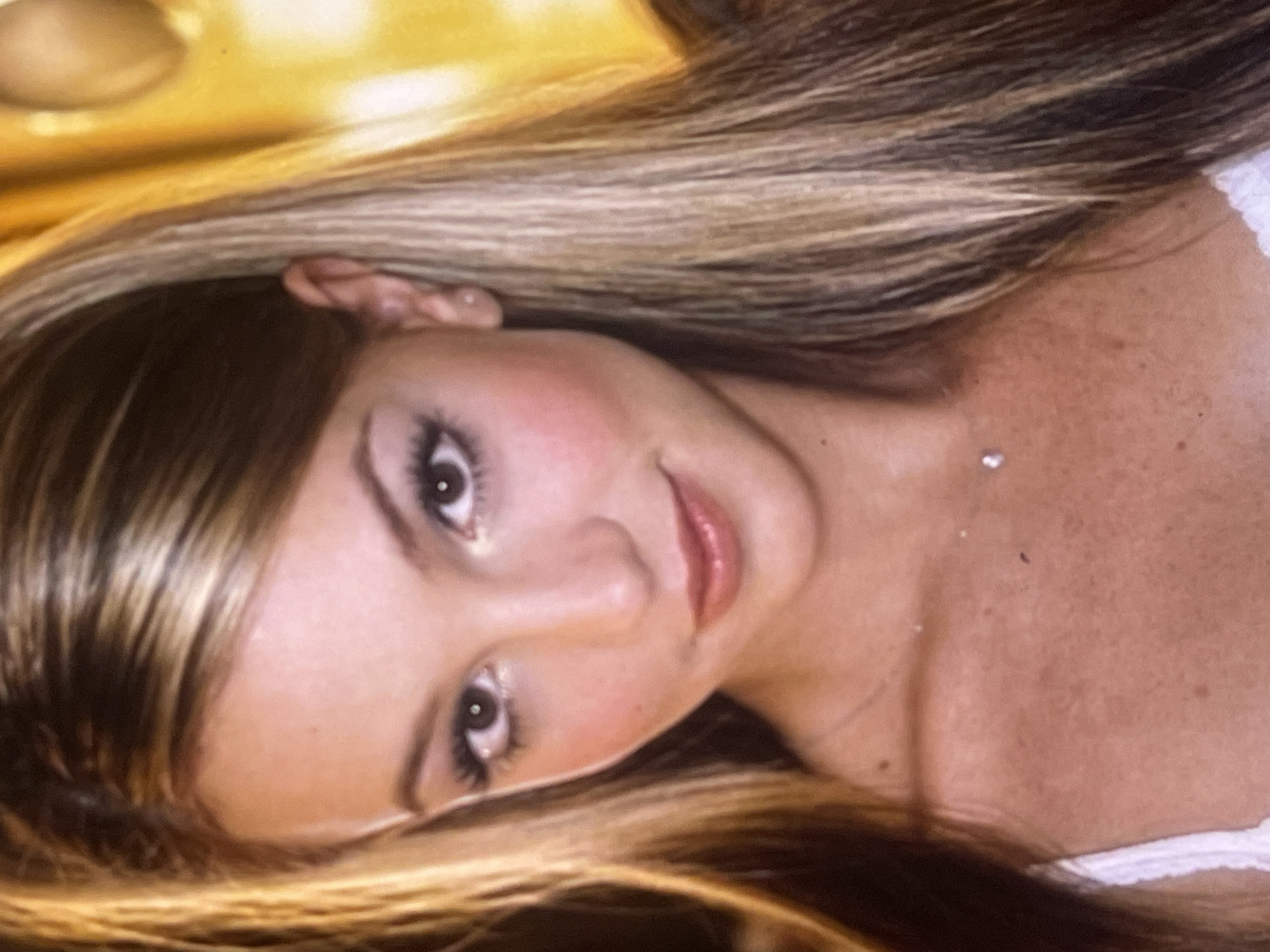 This is a close-up portrait of a young woman, likely taken indoors in what appears to be a personal setting such as a bedroom. The subject is positioned lying down or reclining, with her head tilted towards the camera. She has long, straight to slightly wavy hair with distinct blonde highlights and darker brown lowlights, fanned out around her head. Her face is softly made up with prominent eyelashes, a gentle blush on her cheeks, and glossy lips. Her eyes are dark and looking directly at the viewer. She wears a small, subtle earring in her visible earlobe and a thin necklace with a small, reflective pendant near her collarbone. A hint of a white garment, possibly a lace-trimmed top or undergarment, is visible on her right shoulder area. The background is out of focus, displaying warm, blurred golden and brownish tones, suggesting soft, ambient lighting. The overall mood of the image is intimate and contemplative. There is no visible text. The image does not contain specific visual cues to identify its location as Poltava, Ukraine, beyond the provided context.