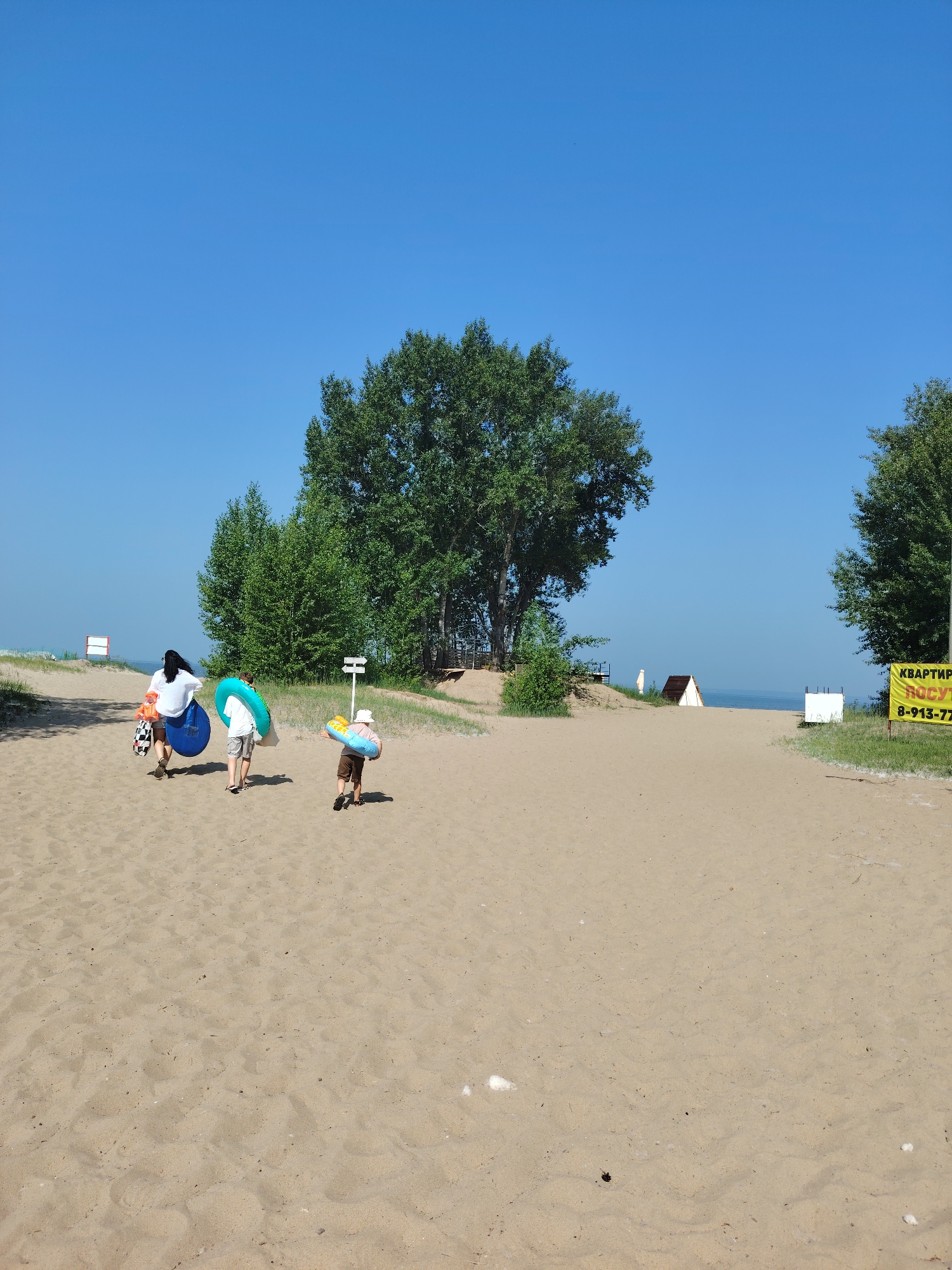A family is walking on a sandy beach on a sunny day. The sky is clear and blue. There are trees on the left and right side of the image, and a large tree in the background. In the distance, there is water. A yellow sign with text in Russian is visible on the right. A family of three, two adults and a child, are walking away from the camera. The adult in the middle is carrying a blue inflatable ring, and the adult on the left is carrying an orange life vest and a blue boogie board. The child in front is wearing a hat and carrying a colorful inflatable ring. They are walking towards a path that leads to the water.