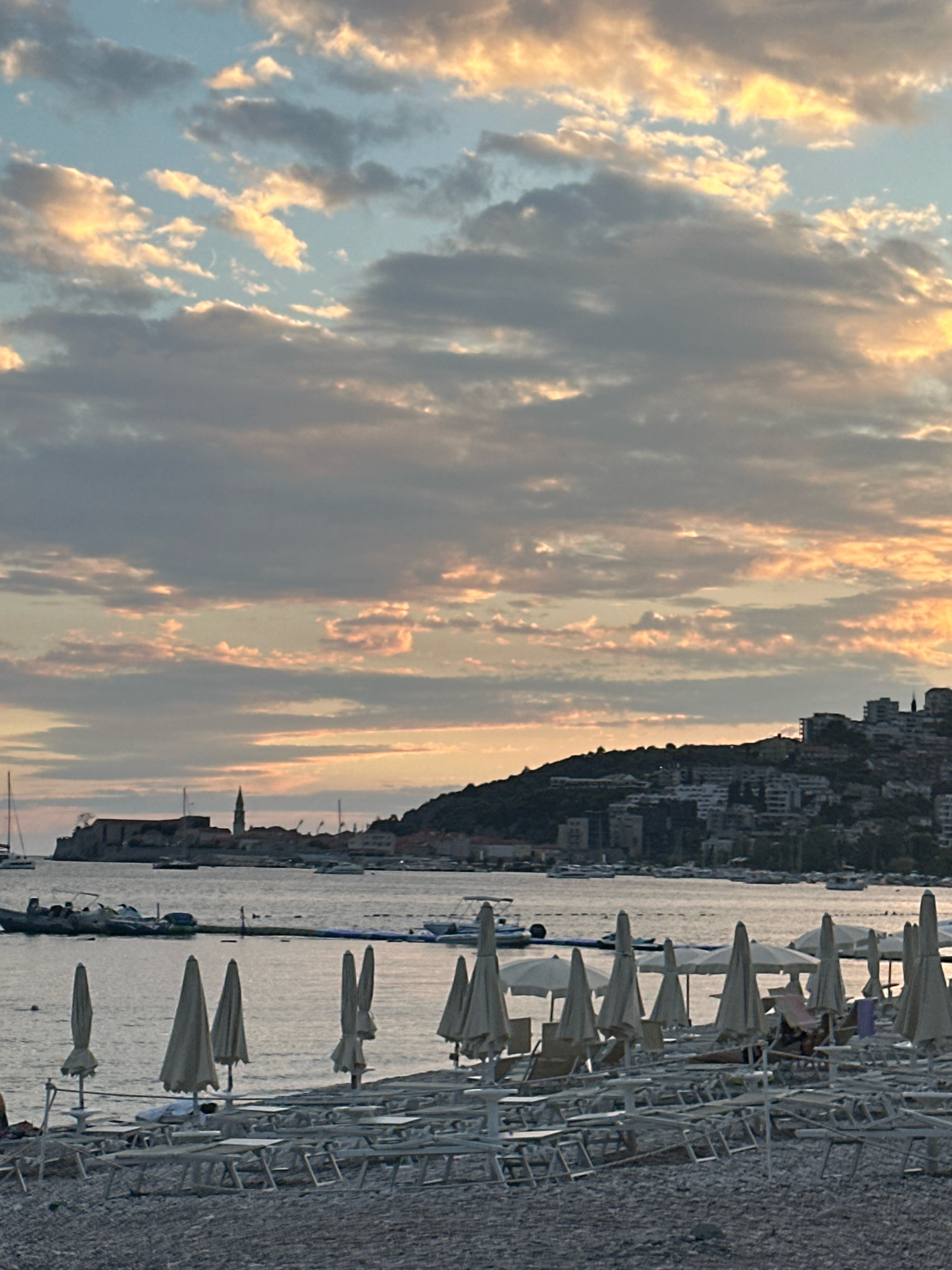 The image depicts a view of the Budva Old Town and coastline at sunset. In the foreground, a pebble beach is lined with numerous empty lounge chairs and closed beach umbrellas, indicating the end of the day. The water is calm, with several boats and yachts anchored in the bay. Across the water, the Old Town of Budva is visible, characterized by its historic stone buildings and a prominent church spire. The background features a hilly coastline dotted with modern buildings. The sky is filled with dramatic clouds, tinged with the warm hues of sunset, casting a soft light over the scene. The overall impression is one of a serene and picturesque coastal evening.