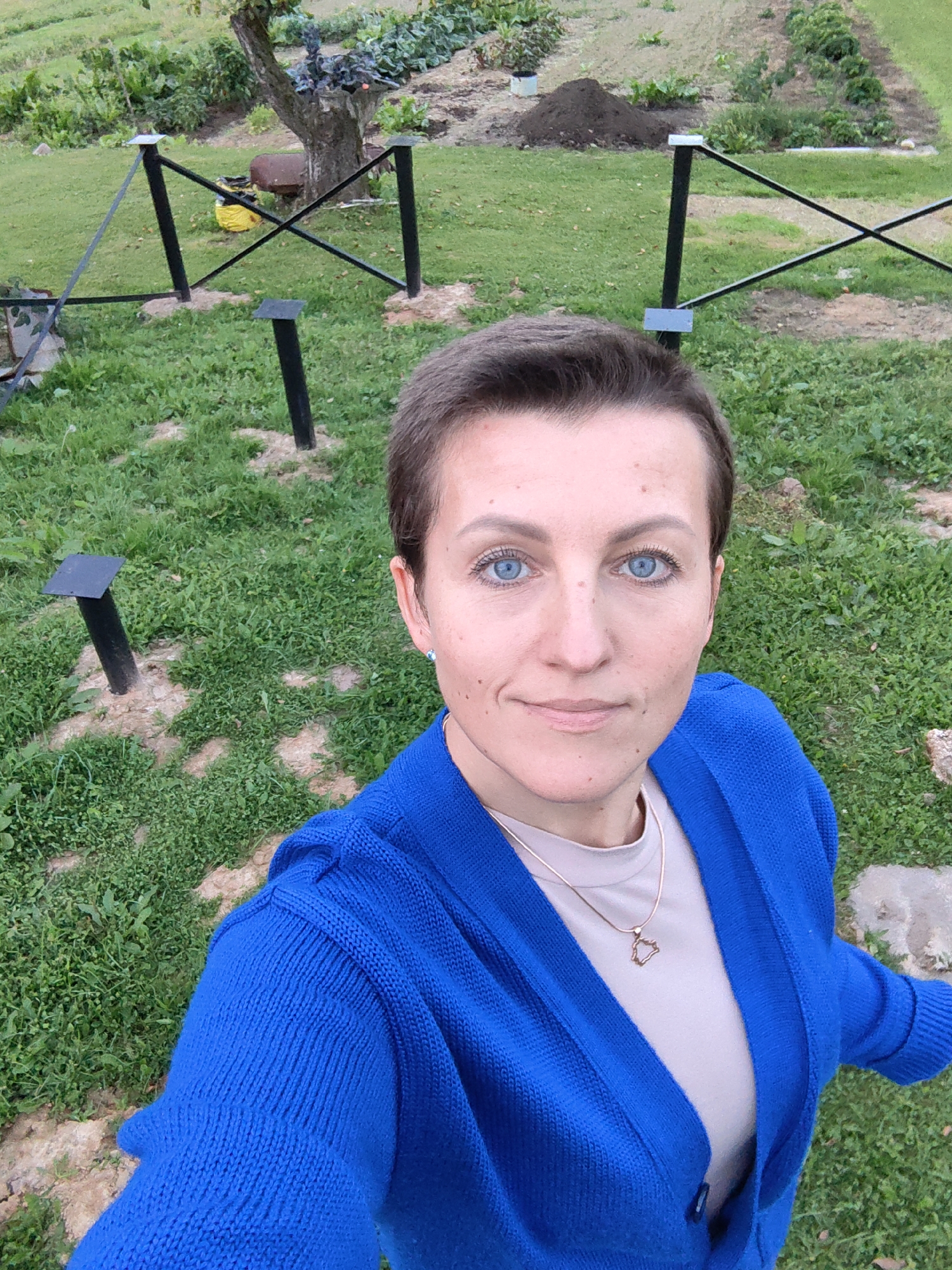 A woman with short brown hair and bright blue eyes takes a selfie outdoors in a grassy area with a garden in the background. She is wearing a bright blue cardigan over a light-colored shirt and a delicate gold necklace. The environment appears to be a rural or garden setting, possibly in the Дзямідавіцкі сельскі Савет, Belarus, given the presence of cultivated rows and natural vegetation. There are several black metal fence posts with square tops visible in the foreground, as well as a tree and a pile of dark soil in the middle ground, suggesting ongoing gardening or landscaping work.