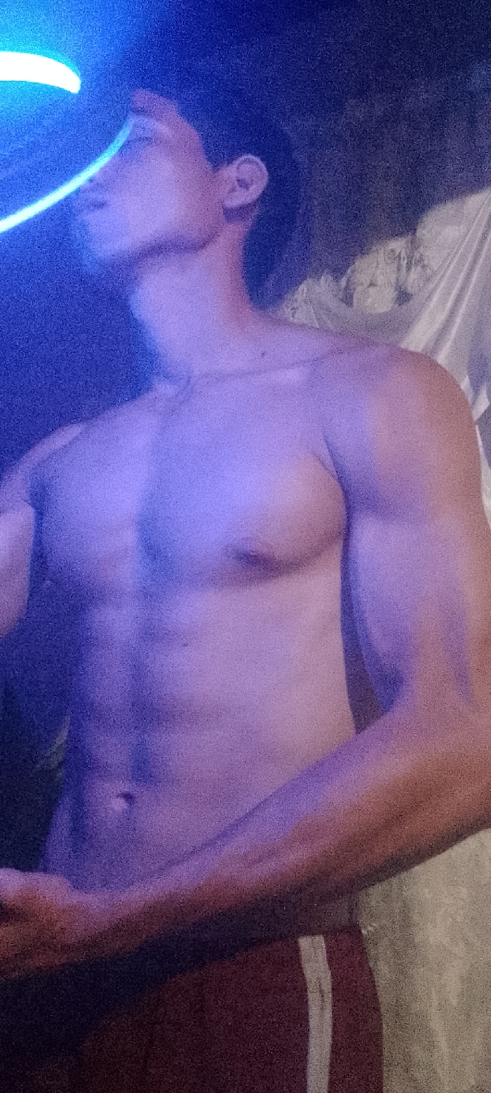 The image displays a male individual, partially nude, captured from the chest upwards, within an indoor or enclosed setting in Juan Griego, Venezuela. The subject possesses a muscular physique, evidenced by defined pectorals, shoulders, and visible abdominal muscles. His head is turned to the left, with his face oriented slightly upwards and partially obscured by shadow and light. A prominent blue light source, possibly an artificial light, illuminates the scene from the upper left, casting a strong blue and purple hue across the subject's skin and the immediate environment. A distinct, bright crescent-shaped light streak is visible in the upper left, adjacent to the subject's head. The background is predominantly dark and indistinct, featuring a light-colored, possibly textured wall or draped fabric on the right side of the frame. The subject is wearing dark red shorts with a single vertical light-colored stripe visible on the right leg. No specific actions or interactions are depicted beyond the subject's pose.