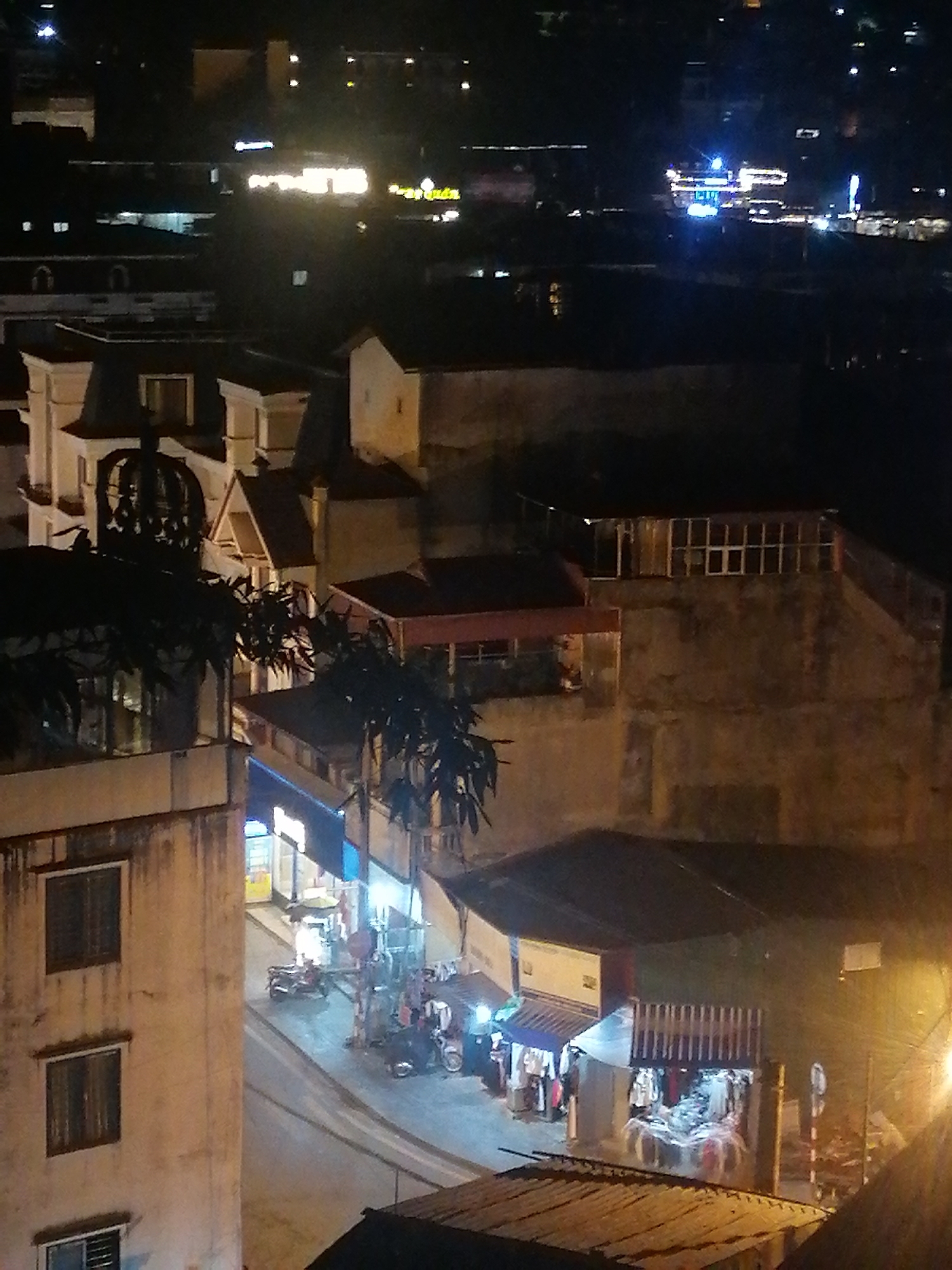 This nighttime shot depicts a street scene in Sa Pa Ward, Vietnam, showing buildings, shops, and a road at night. The image is taken from an elevated perspective. In the foreground, multi-story buildings with windows are visible. Below, the street is illuminated by artificial light from shopfronts and streetlights. Several small shops line the street, some displaying clothing. Scooters are parked along the road. Other buildings of varying heights and architectural styles are in the background. In the distant background, city lights are visible. The overall atmosphere is dark with illuminated areas. No people are visible.
