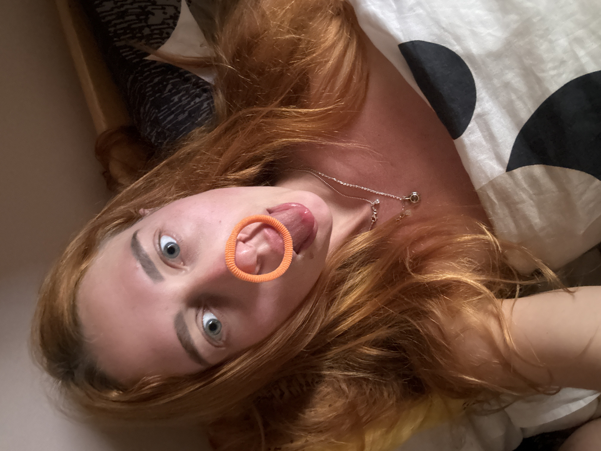 A high-angle portrait captures a fair-skinned female with long, reddish-blonde hair lying in a bed. Her head is tilted slightly to the right, and her wide, light blue eyes gaze directly into the camera. An orange, ribbed hair tie is positioned on the bridge of her nose, partially deforming it, while her tongue is extended outwards from her mouth. She wears two layered silver necklaces, one with a small circular pendant and another with smaller, scattered accents. Her left arm is visible on a yellow pillow to the left. She is partially covered by a white duvet featuring large, black, semi-circular patterns. A plain tan wall is visible in the upper background, along with a darker, patterned fabric to the upper right. While no specific environmental features within the image confirm the location, this scene is described as taking place in Budva, Montenegro.