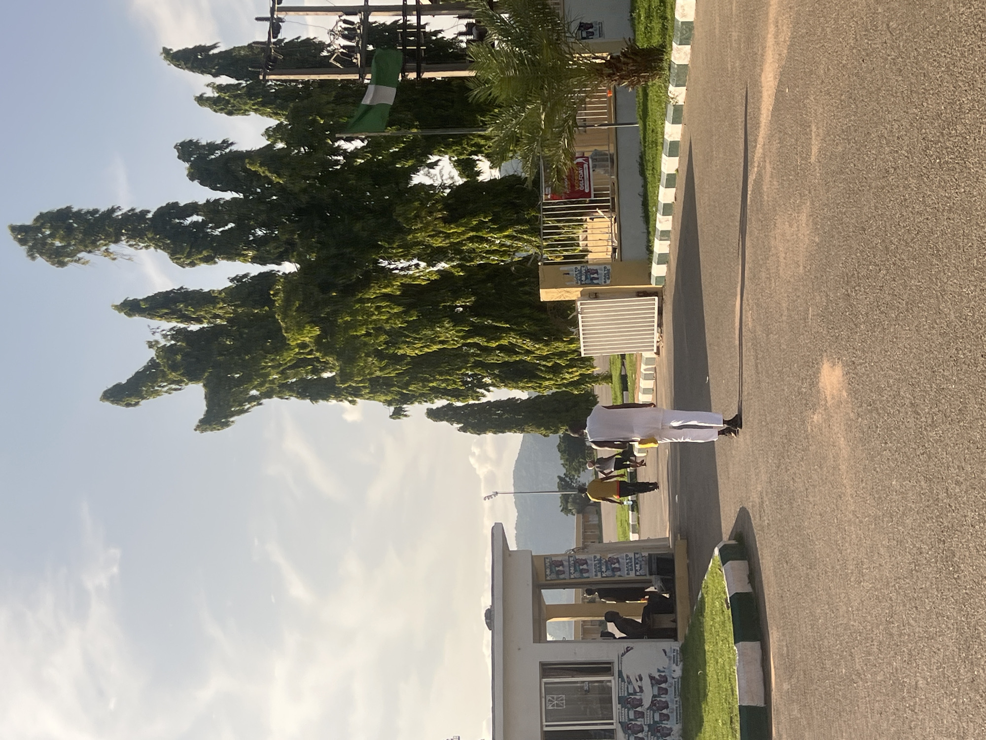 The image portrays an outdoor street scene in Jalingo, Nigeria, bathed in bright daylight. An individual, dressed in white, stands on the right side of an asphalt road, facing away from the viewer and towards the distant horizon. Their long shadow, cast across the road, suggests either early morning or late afternoon.
On the left side of the road, a small, white, flat-roofed building, possibly a guardhouse or kiosk, is visible. Its facade is adorned with several posters, one of which is a red sign with the partially discernible text "GALADIMA." Other indistinct figures can be faintly seen further down the road near this building. A dense line of tall, green trees, along with a solitary palm tree, flanks the left side of the road. A green and white striped Nigerian flag flies from a pole nestled among these trees. The road's edge is clearly marked by a green and white painted curb. Utility poles and overhead wires are visible in the upper left portion of the frame. In the background, a large, tree-covered mountain range rises under a bright blue sky dotted with scattered white clouds.