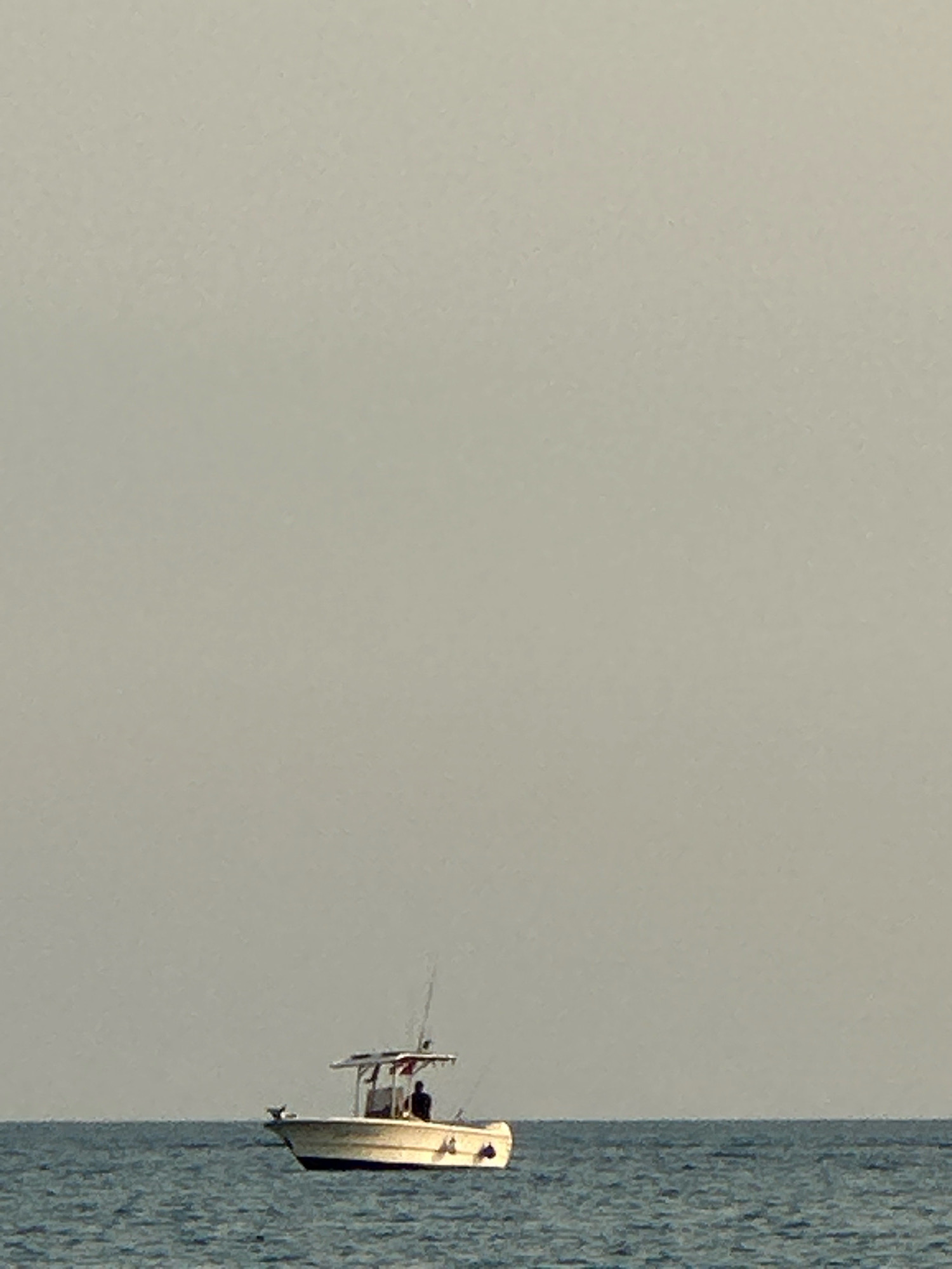 A single fishing boat with a white hull and a canopy is adrift on the calm, deep blue water. The boat has a person seated inside, facing towards the left side of the frame. Two fishing rods are erected from the back of the boat. The sky is a pale, uniform grey, suggesting an overcast or hazy day. The horizon line is clearly defined where the water meets the sky. There are no visible landmasses or structures in the image. The scene appears to be set at sea.