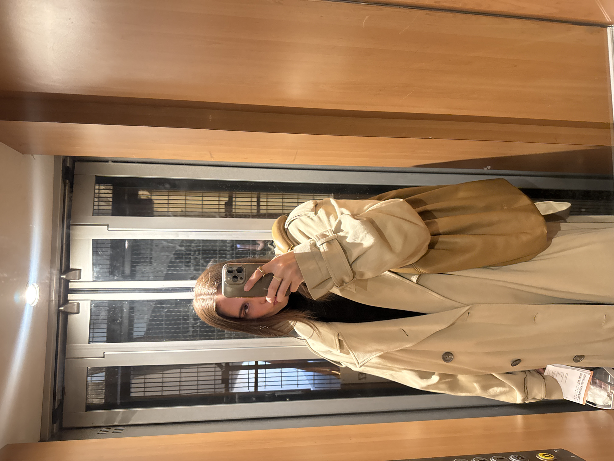 A woman is taking a selfie in what appears to be an elevator. She's wearing a beige trench coat over a black top. Her right hand is holding a mobile phone up to her face. The elevator has wooden paneling on the walls. Through the glass door, buildings can be seen, suggesting the elevator is in a building in an urban area. A document with Russian text is visible in the bottom right. Based on the architectural style visible through the elevator doors and the presence of Russian text on the document, the location might be in Paris, France.