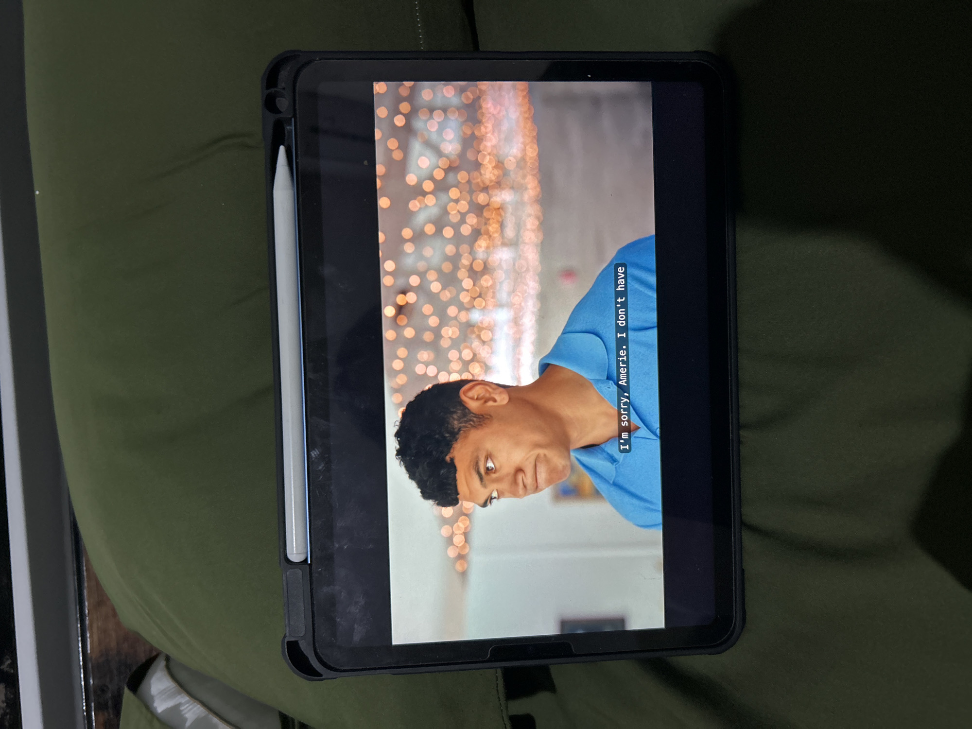 A tablet computer displays a scene from a video, featuring a man with dark, curly hair wearing a blue collared shirt. He is looking off to the left with a concerned expression. Subtitles at the bottom of the screen read, "I'm sorry, Amerie. I don't have". The background of the video is blurred, showing bokeh-like lights. The tablet is placed on a green surface, possibly a bed or a couch, and a stylus is attached to its top edge. The tablet is enclosed in a dark-colored protective case. The scene does not provide any information about the location in Sukawati, Indonesia.