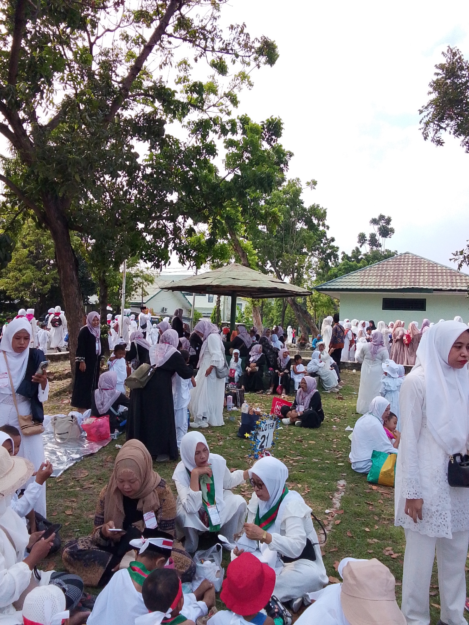 The image depicts a gathering of people, predominantly women and children, dressed in white Islamic attire, in an outdoor setting. The scene appears to be a park or communal area with grass, trees, and a small building with a tiled roof in the background. Several individuals are seated on the ground, some on mats or cloths, while others are standing or walking. Many are wearing hijabs, and some children have small red and white hats. The overall atmosphere suggests a religious or community event, possibly related to Eid or a similar celebration. Based on the distinctive architectural style of the building with its prominent green roof tiles and the style of dress, it is likely located in Cirebon, Indonesia. The presence of picnic mats and scattered leaves on the ground indicates a relaxed, daytime outdoor gathering.