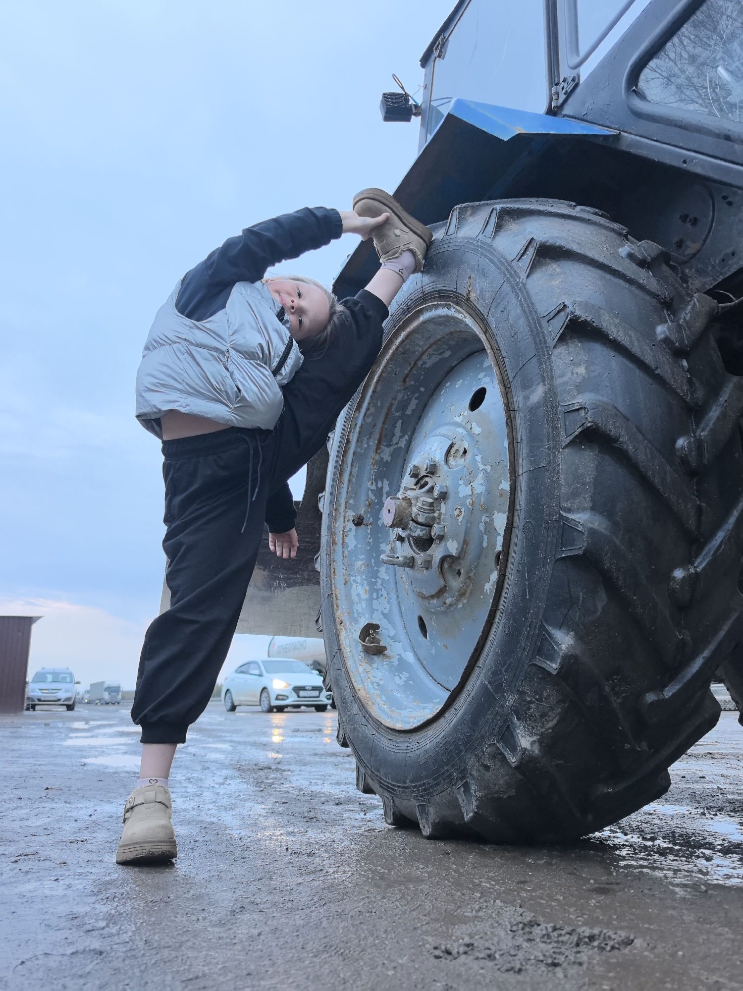 A young girl is performing a split against the tire of a blue tractor. She is wearing black sweatpants, a black jacket, a silver puffer vest, and tan boots. The tractor tire is large and heavily treaded with a light blue rim. The ground is wet, and several cars are visible in the background. The scene is set in Bol'shoi Mashlyak, Russia.