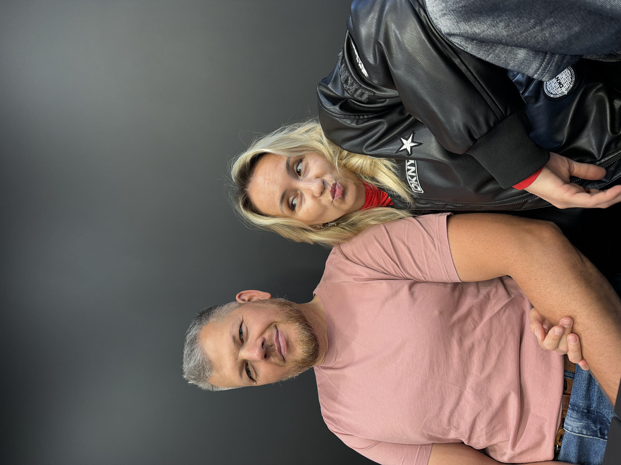 The image features two people, a woman and a man, against a plain gray background. The woman, with blonde hair, is positioned behind and above the man, leaning in towards the camera with her head tilted. She is wearing a black leather jacket with a white star and "DKNY" logo on it, and a red top underneath. She has a playful expression, puckering her lips. The man, with short gray hair and a beard, is in the foreground, looking towards the camera with a slight smile. He is wearing a pink t-shirt and blue jeans. Their arms are clasped behind their backs. The lighting is even, suggesting an indoor setting. There is no indication of the time of day, weather, or specific location beyond the general context of Izhevsk, Russia. The overall mood appears to be informal and friendly.