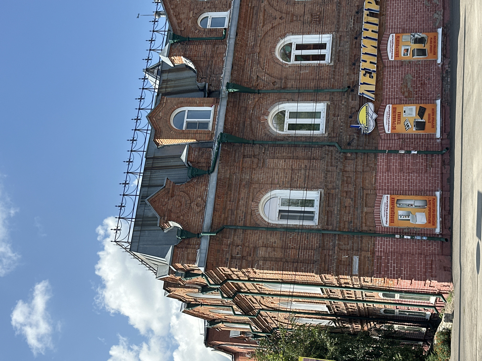 The image shows the brick facade of a historic building with an arched mansard roof, located in the city of Biysk, Russia. The building has multiple arched windows with white frames and is decorated with green downspouts. A large sign with the name "ЛЕНИНГРАДСКИЙ" (Leningradsky) in yellow letters is visible on the facade, indicating a commercial establishment. Below the main sign, there are three vertical posters advertising various household appliances such as refrigerators, ovens, and stovetops. The building is situated on a street with a paved road and sidewalk in the foreground. The sky above is blue with scattered white clouds. The overall scene depicts a well-preserved architectural structure within an urban environment.