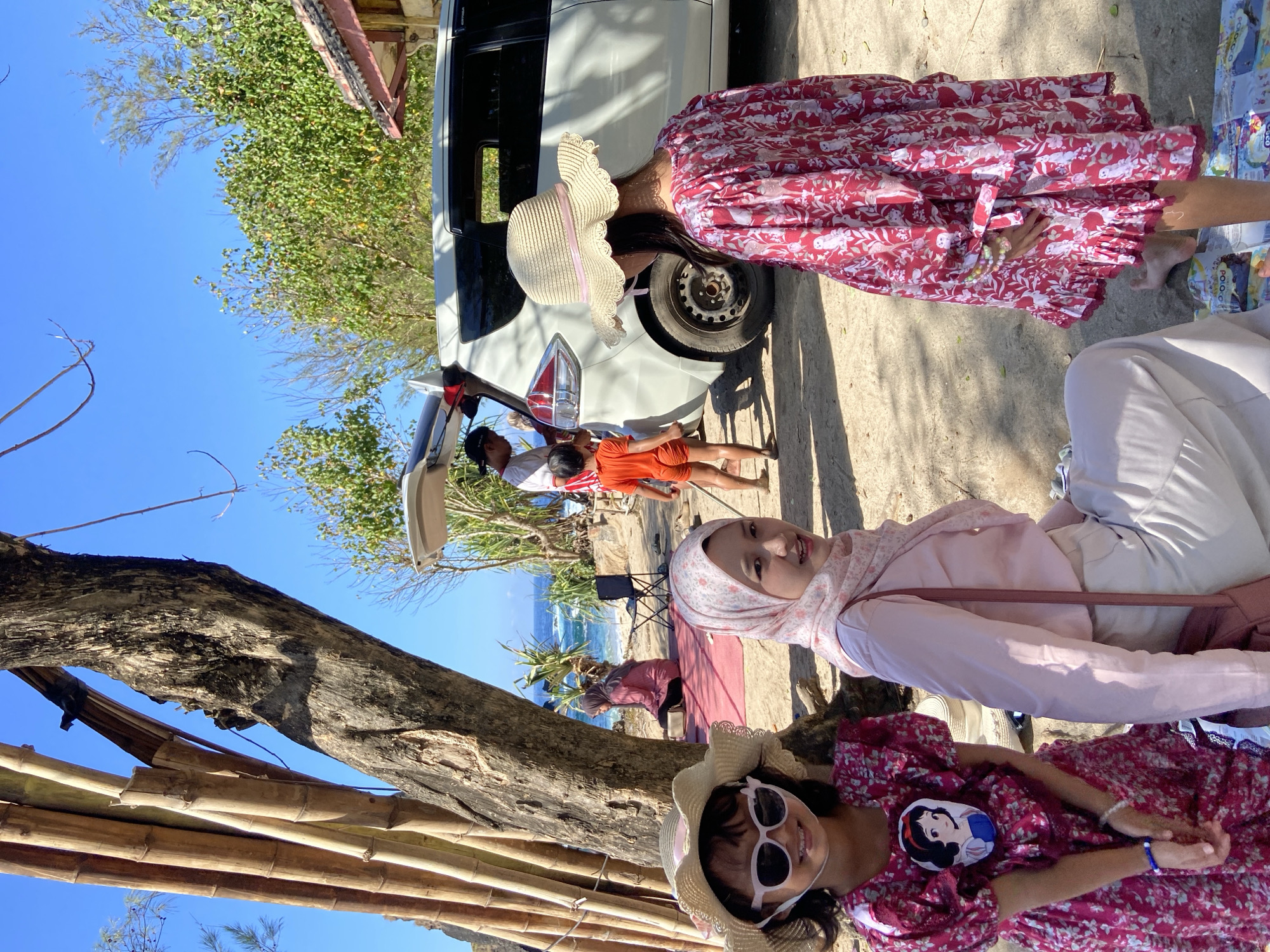 The image depicts a family gathering near a beach in an unknown location in Indonesia. A white vehicle is parked nearby, with the trunk open and a man and a child visible inside. In the foreground, two females wearing hats and dresses are relaxing near a bamboo structure. Another female in a hijab is also present. The background shows the ocean and some vegetation.