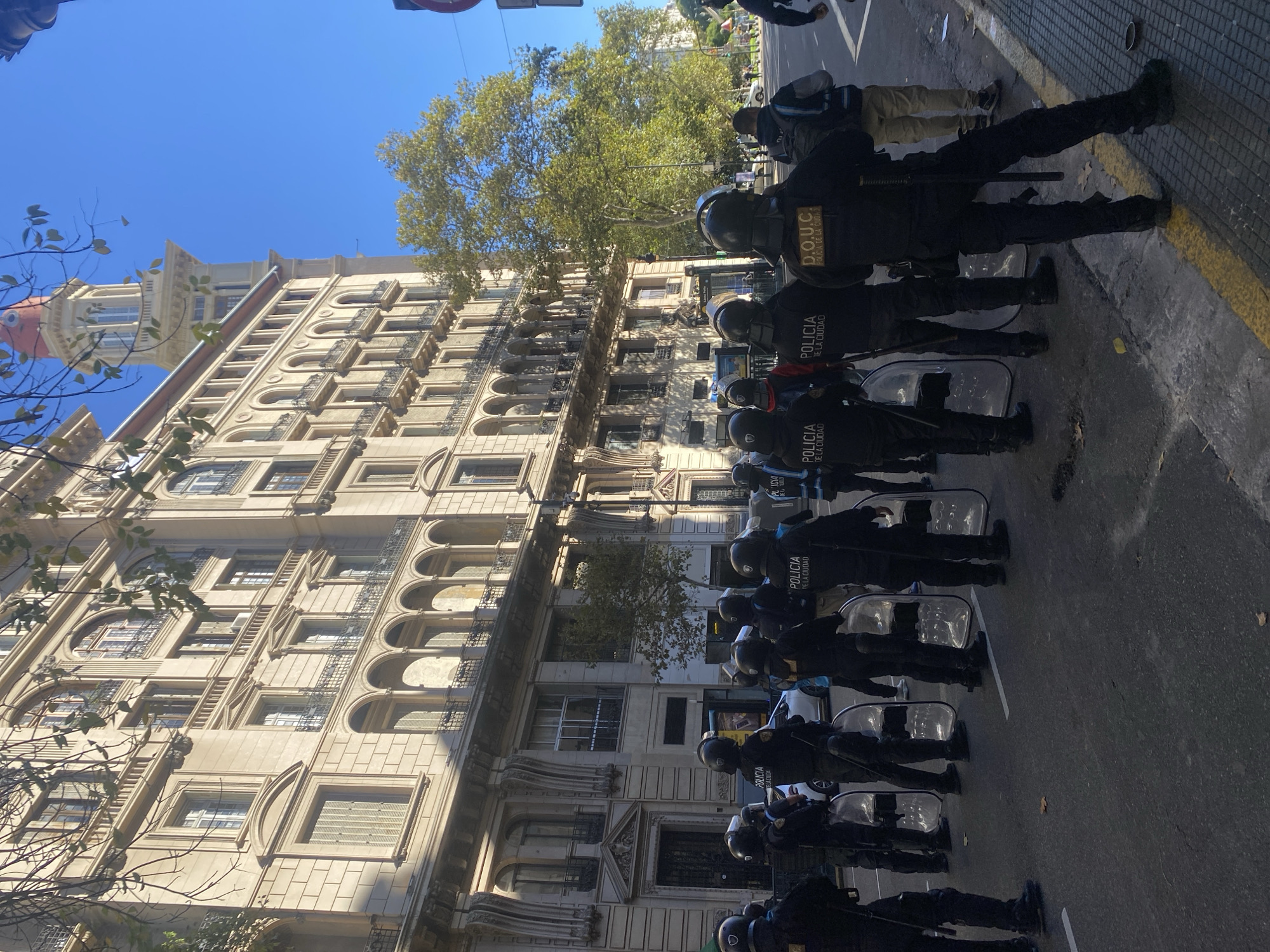 The image depicts a daytime street scene in an urban area, likely Buenos Aires, Argentina, under a clear blue sky. A line of approximately a dozen police officers, equipped in full riot gear, stands in a row facing forward (if the image were rotated correctly). Each officer wears a dark uniform, a protective helmet, and a vest. Many are holding clear rectangular riot shields and carrying batons.
Visible text on the backs of the officers' vests reads "POLICIA DE LA CIUDAD" (Police of the City), confirming their affiliation with the Buenos Aires City Police. One officer's vest also displays the acronym "D.O.U.C.", which likely refers to a specialized unit within the police force. They are positioned along a paved street or sidewalk, distinguished by a yellow painted line.
To their left (if the image were correctly oriented) stands a grand, multi-story building featuring classical European-style architecture. The light-colored facade includes numerous arched windows, some with decorative iron balconies or grilles, and intricate stone detailing, characteristic of historic structures found in Buenos Aires. Green trees with full foliage are visible behind and above the building, indicating a pleasant, sunny day. The scene suggests a prepared police presence, potentially for crowd control or security during an event.