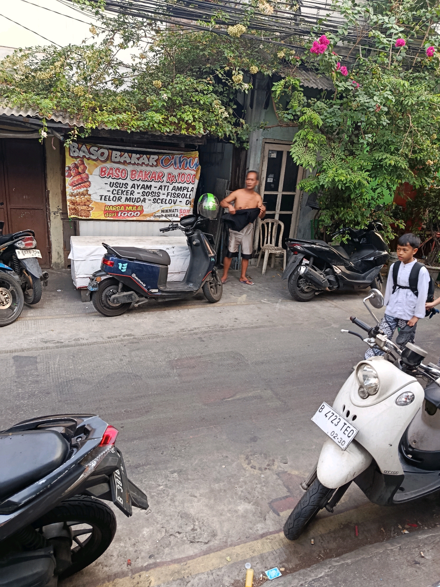 The image shows a street scene in North Jakarta, Indonesia, with several motorcycles parked along the side of a building. A man is standing shirtless in front of the building, holding a piece of dark fabric. A young boy wearing a backpack stands near a white scooter. A banner advertising "Baso Bakar" (grilled meatballs) is visible on the building. Green vines and pink flowers are growing around the structure. The street appears to be paved with asphalt and shows signs of wear and tear.