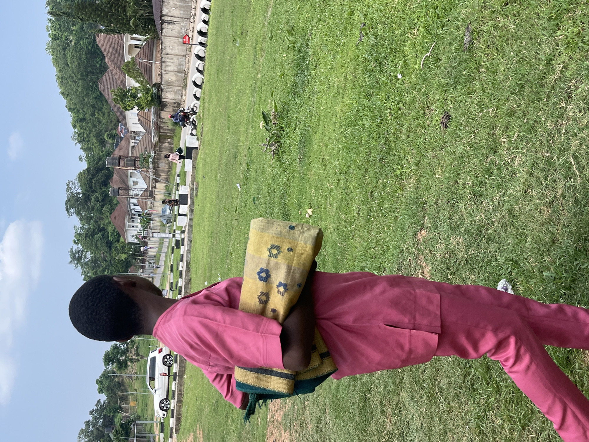 A person stands in a grassy field, looking to the left. They are wearing a pink outfit, with a yellow and blue patterned cloth wrapped around their arms. The person's head is turned away from the camera, showing the back of their head. In the background, there are houses with brown roofs, a road with white and black barriers, and some trees. A white car is parked on the road. The sky is blue with some clouds. The scene appears to be outdoors, likely during the daytime. The overall setting suggests a residential area in Abuja, Nigeria, with a mix of modern housing and green spaces. There are no visible text elements in the image.