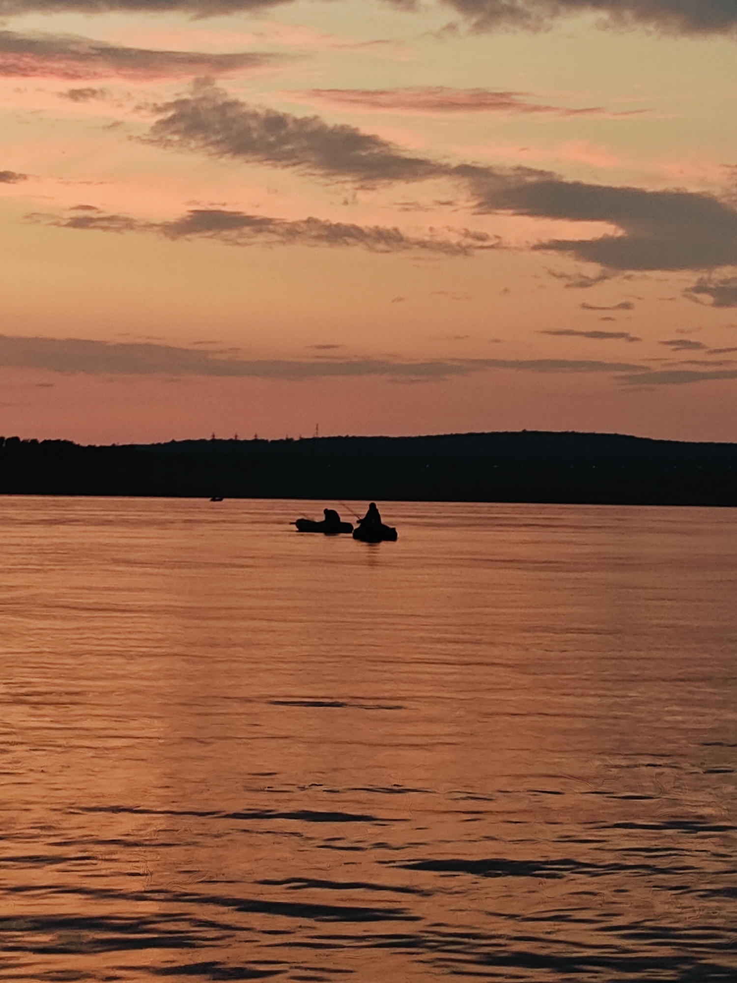 At sunset, two people are fishing in separate boats on the Kama River in Chaikovsky, Russia. The sky is filled with streaks of pink and orange clouds. The water reflects the colors of the sky, creating a warm, golden hue. In the distance, a dark, silhouetted treeline marks the shore. The overall scene is tranquil and serene.