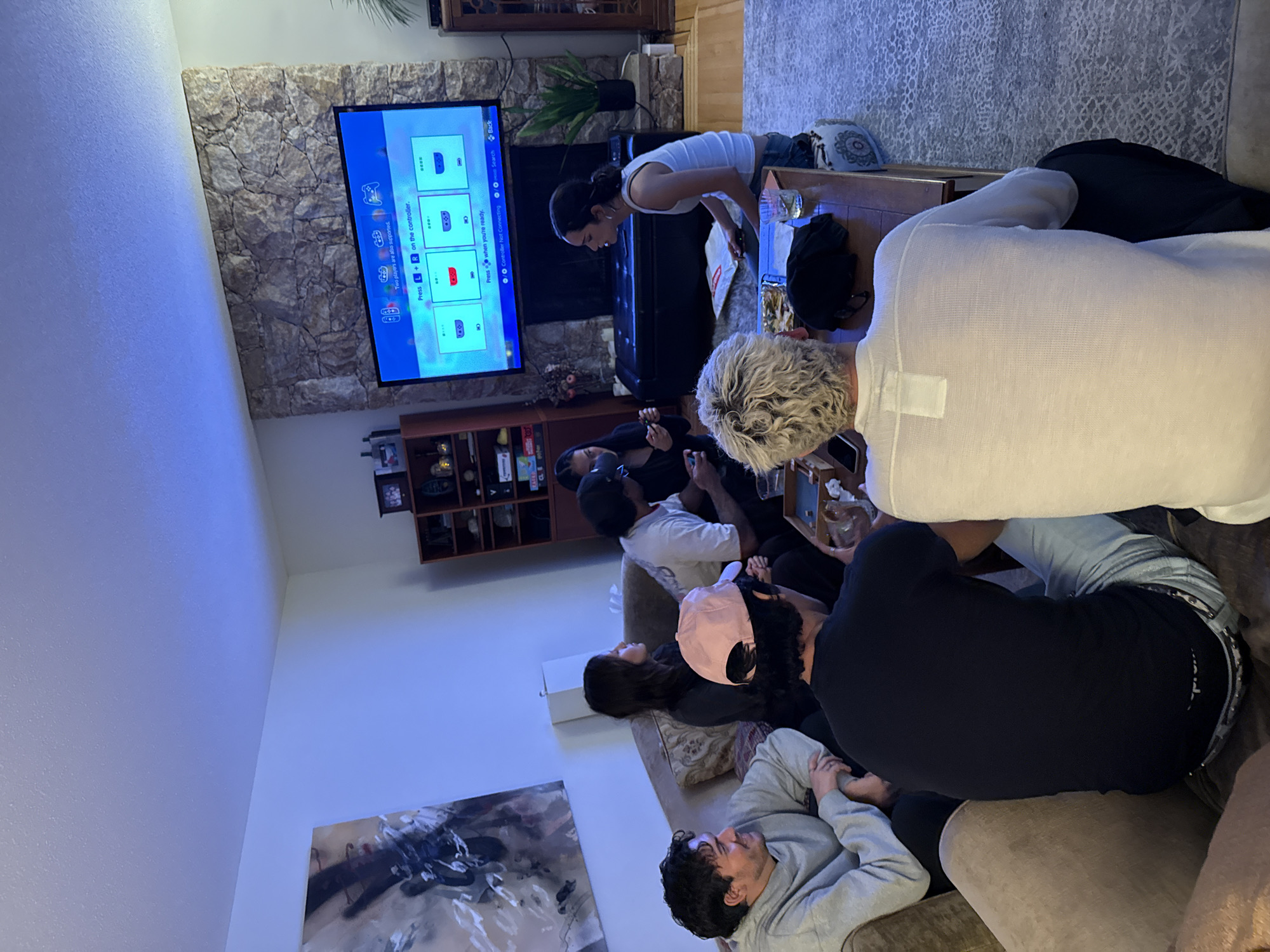 A gathering of people is present in a living room setting. Approximately eight individuals are either sitting or gathered around a central coffee table and watching a television mounted on a stone wall. The television displays what appears to be a game setup screen. A woman is leaning over the coffee table, perhaps arranging items on it. Seated on couches, the other individuals appear to be focused on the television. The location is in District of North Vancouver, Canada.
