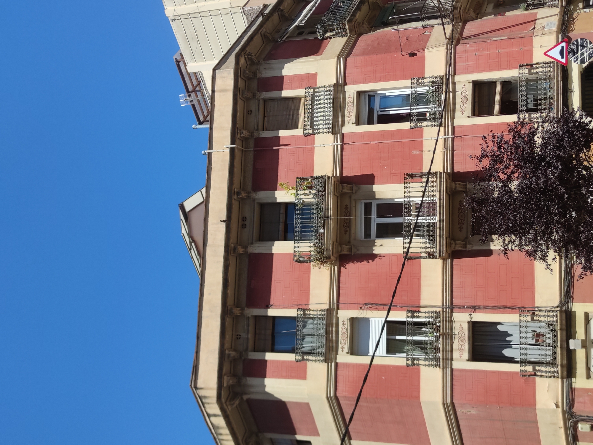 The image shows a building with a red and beige facade, adorned with ornate metal window grilles. The sky is a clear blue. A tree with dark foliage is visible on the right side of the image. A triangular street sign is positioned near the top right corner. The location is Unknown, Unknown.