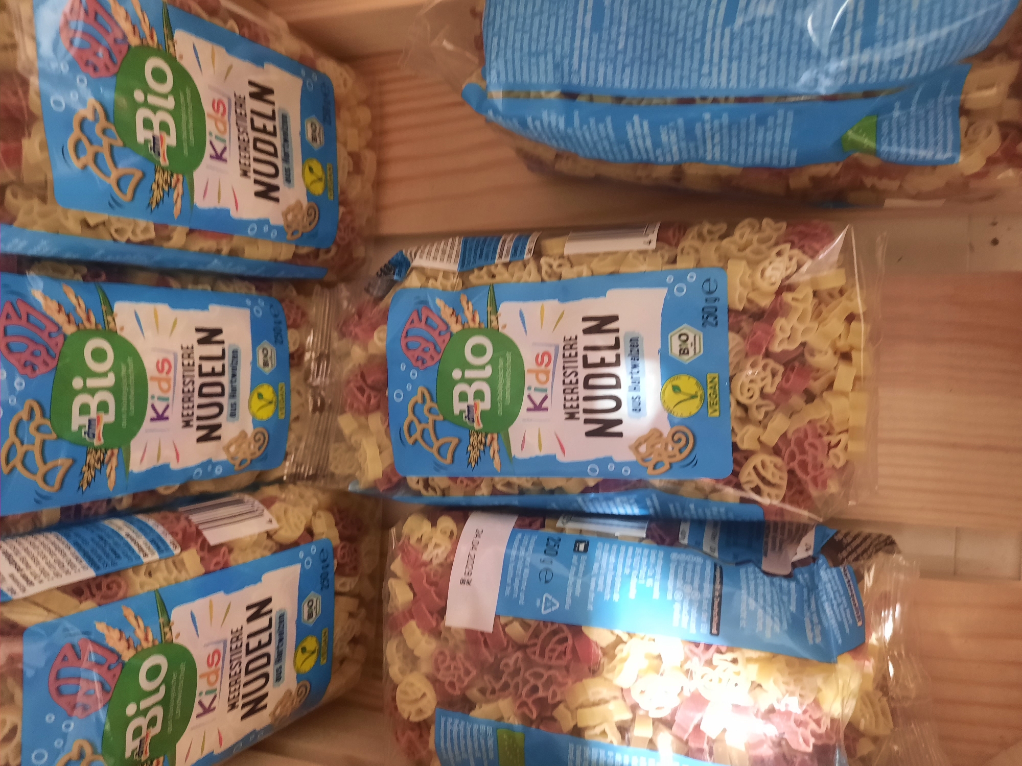 The image shows several bags of "Bio Kids Meerestiere Nudeln" (organic kids' sea creature pasta). The bags are blue with illustrations of sea creatures and wheat. The pasta inside is shaped like various sea animals and shells, in yellow and pink colors. The bags are arranged in a wooden crate. One bag has a label with "24. 04. 2024" which may indicate a date. The scene is lit from above, creating shadows within the crate. The image does not provide any information about the city of Rosenheim, Germany.