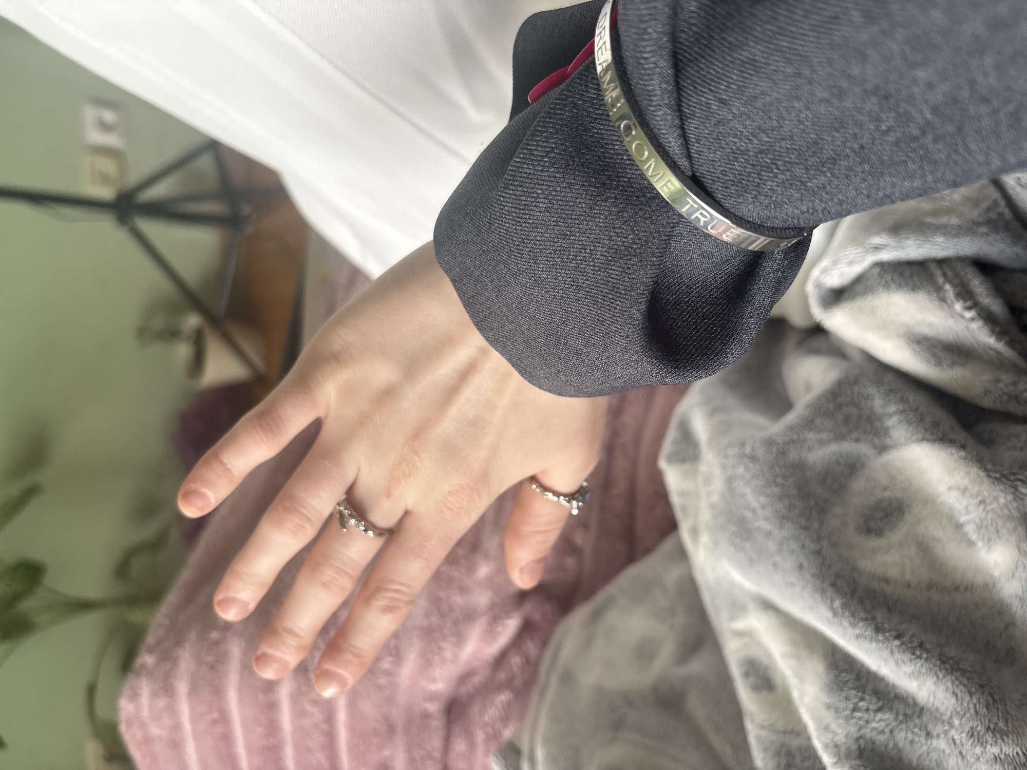 The image captures a close-up of a person's left hand and lower arm, positioned over soft textiles in an indoor setting. The hand, with light skin tone, features manicured nails and wears two silver-colored rings with intricate, possibly stone-set designs on the middle and ring fingers. The arm is clad in a dark grey long-sleeved garment, appearing to be a jacket or blazer, beneath which a white garment is visible. Around the wrist, a silver bangle bracelet is worn, engraved with text.
The primary activity depicted is the resting of the hand and arm. The background suggests a cozy, personal environment. To the left, a blurry light green wall is visible, with a black metal stand (possibly a tripod) and electrical outlets in the distance. Below the hand, a soft, light pink fabric, possibly a blanket or clothing, creates a gentle texture. To the right, a plush grey and white patterned blanket adds another layer of comfort to the scene.
A notable detail is the text engraved on the silver bracelet, which reads "DREAMS COME TRUE", with "DREAMS!" and "COME TRUE" clearly visible from different angles. The varying textures of the fabrics – the fine weave of the dark grey sleeve, the smooth white material, the soft pink textile, and the fluffy grey and white blanket – contribute to the tactile feel of the image. The lighting is soft and even, typical of an indoor setting, without indicating a specific time of day or particular weather. No explicit emotions are conveyed, presenting a calm and composed scene.