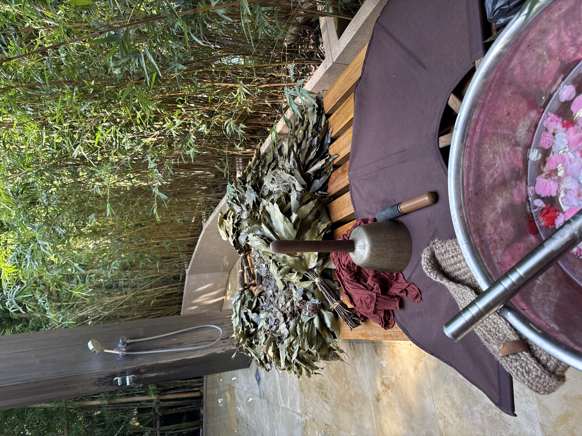 The image shows an outdoor spa setting. Bamboo forms a backdrop. A wooden bench holds sauna accessories: bundles of dried leaves, a metal bowl filled with water and flower petals, a small mallet, a brown cloth, and a scrub mitt. A showerhead is visible on the lower left. The location is Kediri, Indonesia.