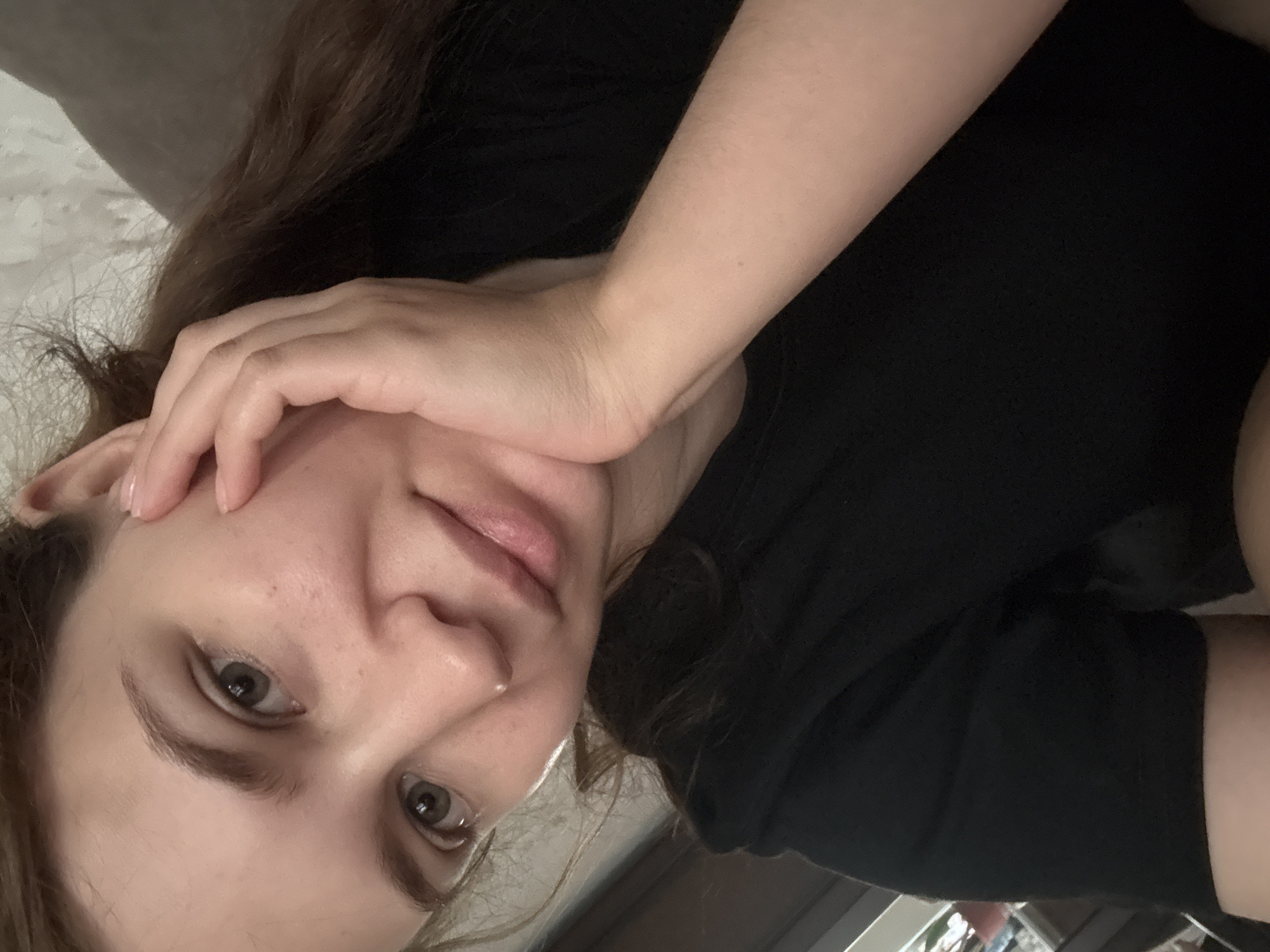 A young woman with light brown hair is shown in a close-up selfie. She is wearing a black t-shirt and resting her chin on her hand. Her skin is fair, and she has light brown eyes. The background is blurred, but appears to be indoors, possibly a living room or bedroom. There is no indication of the location being in Chaikovsky, Russia.
