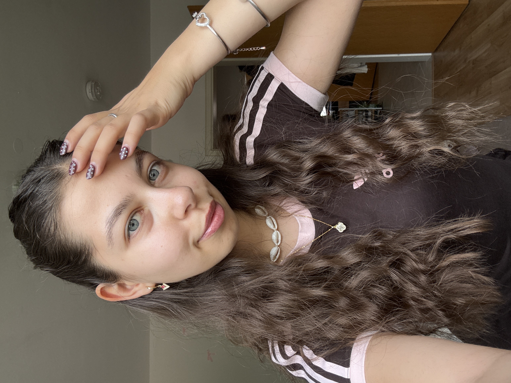 A young woman with long, wavy brown hair is posing for a selfie indoors. She is wearing a dark brown Adidas-style shirt with light pink stripes and trim. She has on jewelry, including a shell necklace, a pendant necklace, a silver bracelet with a heart charm, and earrings. Her nails are painted dark with white dots. Her right hand is raised, with fingers touching her forehead. Her expression is serene.
The background is partially obscured, showing a doorway leading to another room with light-colored walls and flooring. An overhead light fixture is visible on the ceiling behind her. The location is likely an interior space in Bruchköbel, Germany.