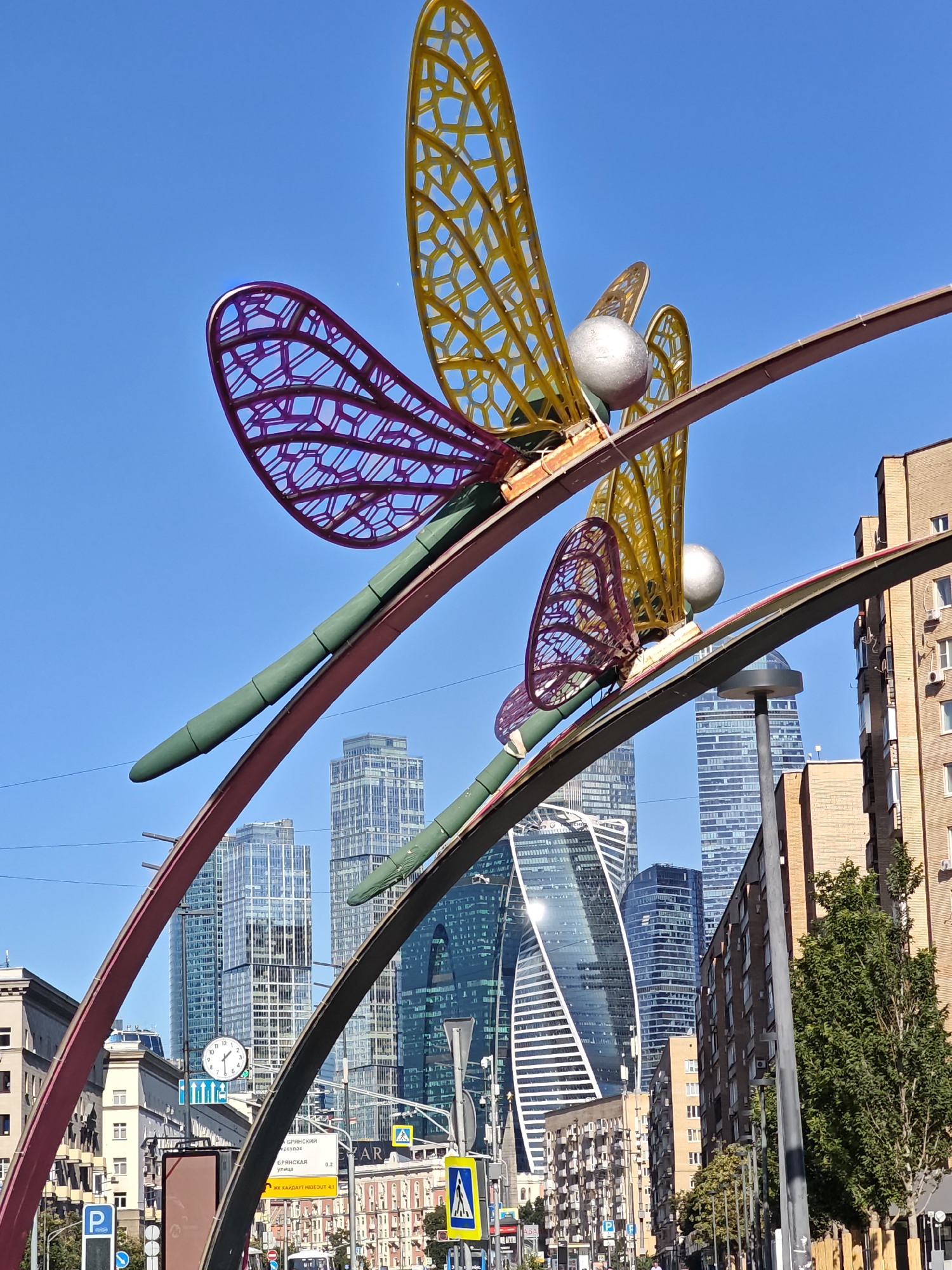 This is an outdoor, eye-level, full-shot photograph taken on a clear, sunny day in Moscow, Russia, likely in the Moscow City district. The foreground features a large, colorful art installation of two dragonflies, constructed from metal. The dragonflies are positioned on curved, burgundy-colored beams, with their wings extended. The larger dragonfly has yellow, honeycomb-patterned wings, a silver sphere for its head, and a green body. The smaller dragonfly has purple, honeycomb-patterned wings, also with a silver sphere head and a green body.
In the background, a cluster of modern skyscrapers rises against a bright blue sky. These buildings are characterized by their sleek, glass facades, with some featuring distinctive geometric designs, such as the twisting "Moscow Towers" (Neva Towers). A few older, brick residential buildings are visible on the right and left sides of the frame, interspersed with the contemporary architecture. Street-level details include traffic signs, streetlights, and a clock mounted on a pole. The presence of these elements, particularly the modern skyscrapers of Moscow City, strongly indicates the location.