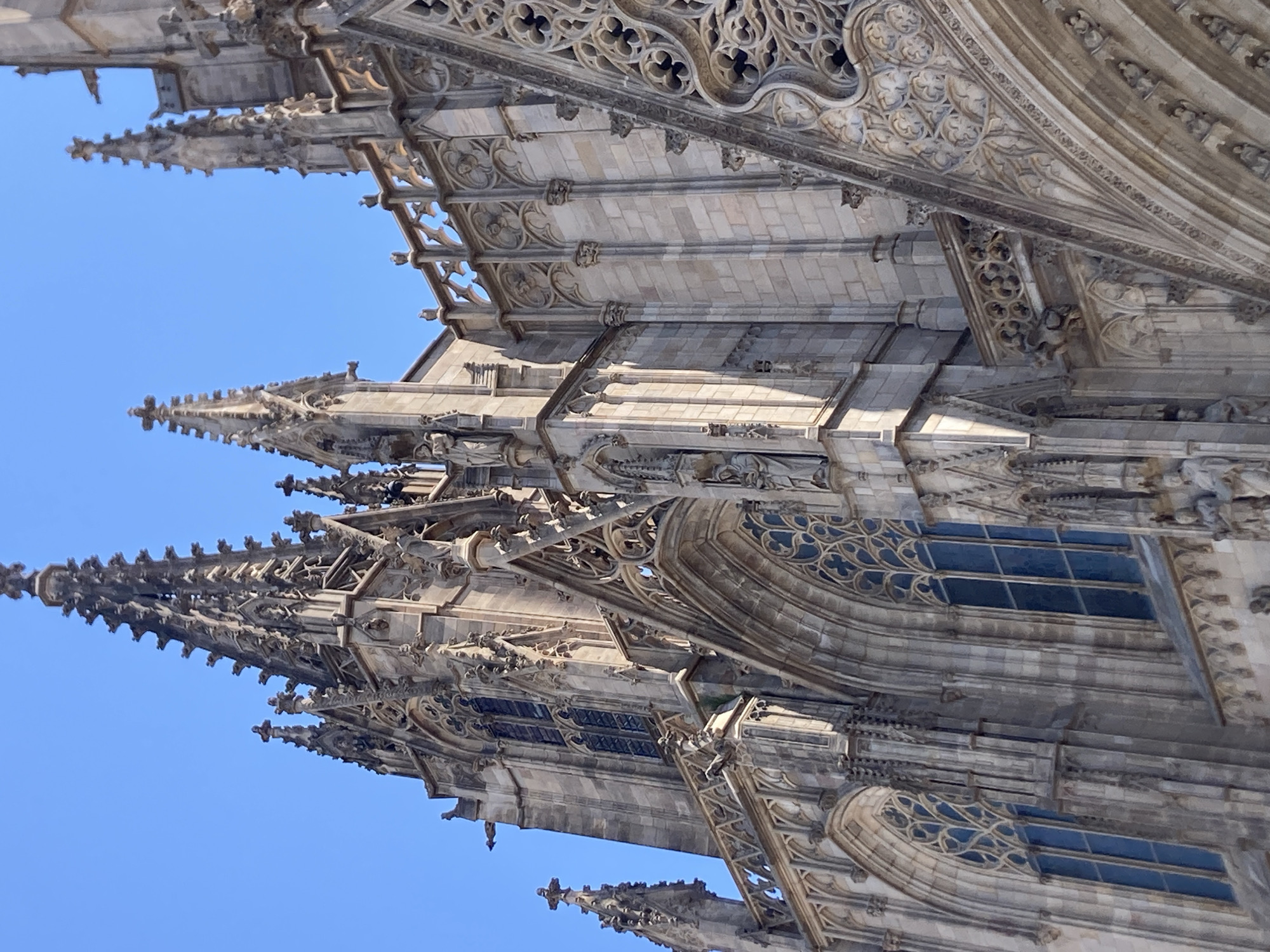 The image shows a detailed, low-angle view of the exterior of the Barcelona Cathedral, located in the Gothic Quarter of Barcelona, Spain. The architecture is Gothic, characterized by tall spires, intricate stonework, pointed arches, and detailed gargoyles. Several stained-glass windows are visible, some with blue panes. The sky is a clear, bright blue. The sunlight casts shadows on the stone facade, highlighting the textures and carvings. There are no people visible in the frame. The focus is entirely on the magnificent and ornate details of the cathedral's upper structure.