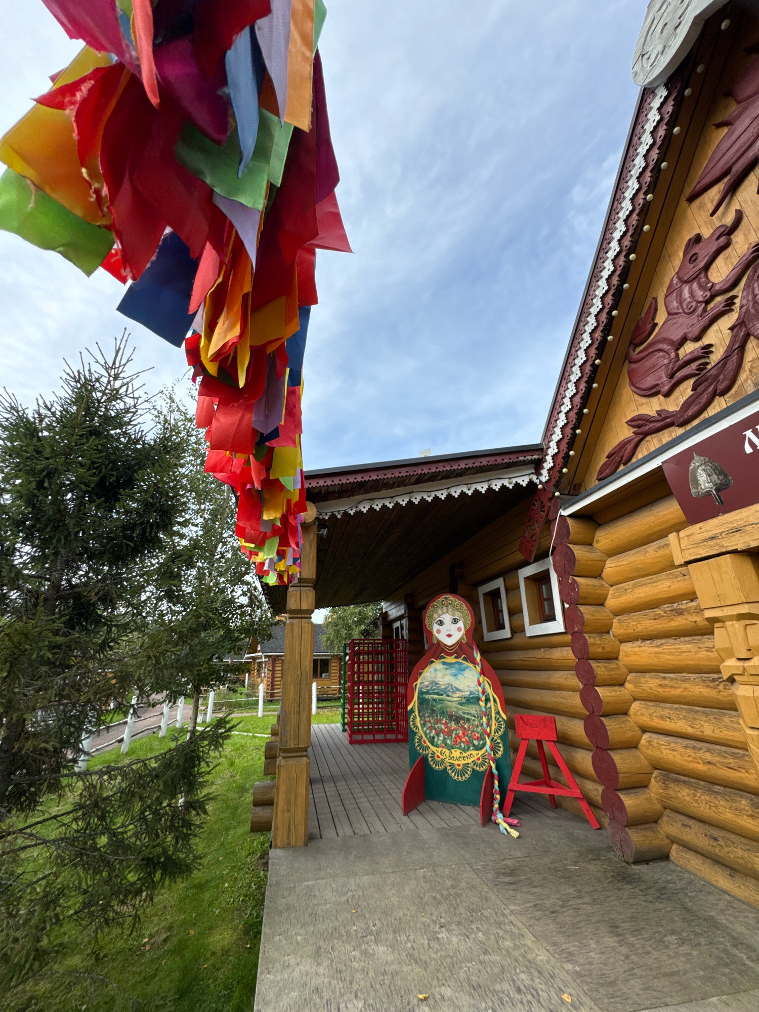 The image shows a traditional Russian wooden house in the city of Peterhof, Russia. In the foreground, a colorful string of fabric flags hangs overhead, framing a wooden porch. A large, flat cut-out of a Matryoshka doll stands on the porch, facing left. Next to the Matryoshka doll, a small, red, A-frame stool is placed on the wooden planks of the porch. The house itself is constructed from large, rounded logs and features decorative carved wooden elements along the roofline and above a window. The roof is covered with dark material, and white ornamental trim with small circular details runs along the eaves. To the left of the porch, a tall evergreen tree is visible, with lush green grass covering the ground. In the background, another wooden building can be seen, along with a red metal fence. The sky is cloudy, with patches of blue visible.