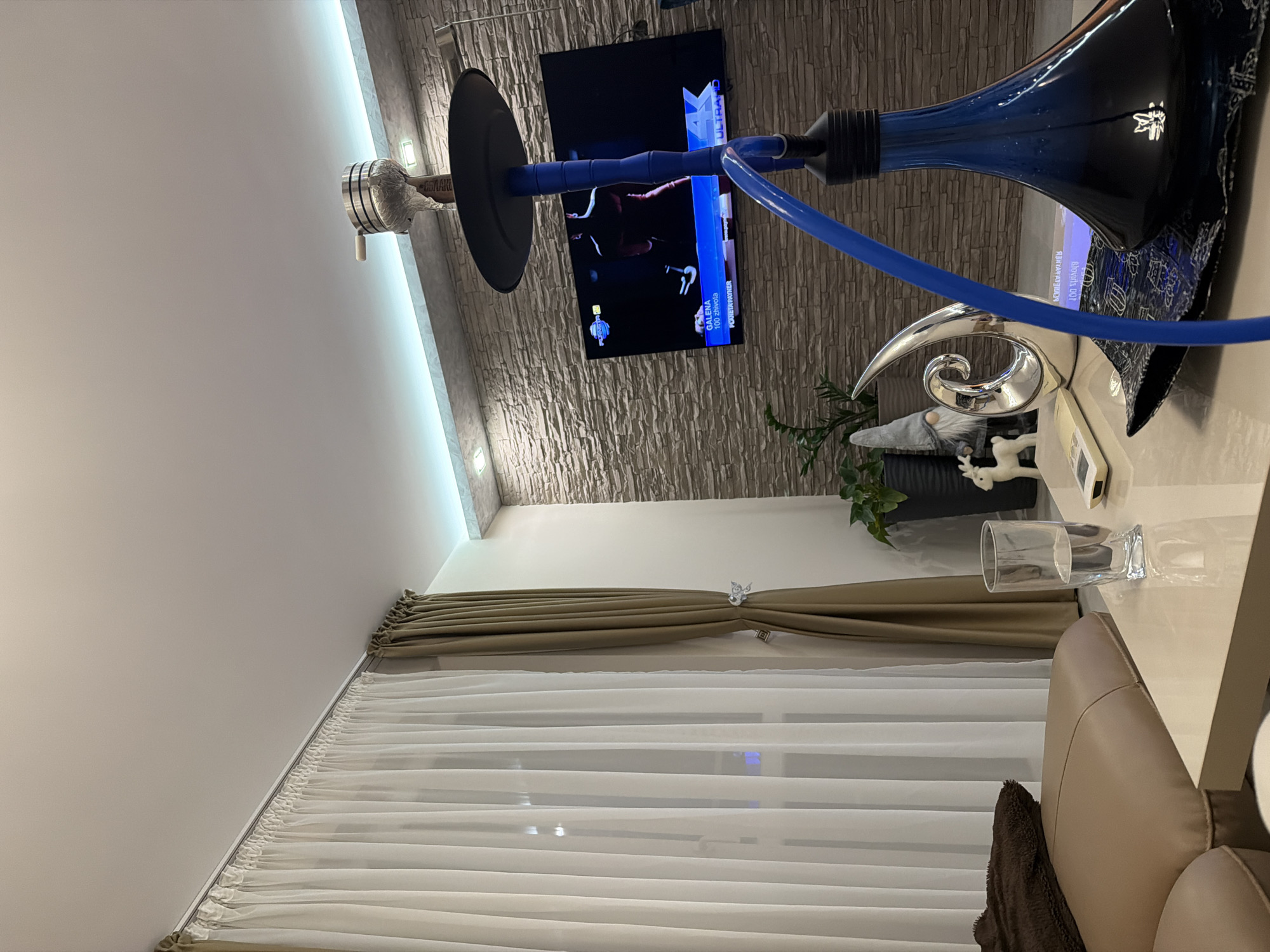 The image displays an interior living space, likely a lounge or living room, seen from a high angle looking downwards and across the room. The room features a modern aesthetic with various decorative and functional elements.
In the foreground, a prominent blue hookah with a matching hose and a black bowl sits on a light-colored table or counter. Next to it, a silver, abstract, swirling sculpture, a small grey gnome figurine, and a white reindeer figurine are visible. A clear glass of water and a remote control also rest on the surface. Behind these items, a potted plant adds a touch of greenery.
The wall behind the table is covered in a textured, stone-like material, possibly slate or stacked stone, giving it a rustic yet contemporary feel. Mounted on this wall is a flat-screen television displaying content that appears to be a sports broadcast or gaming, with visible text including "ULTRA", "GALENA", "100 EUROSA", and "POBETAVPARTNER". Small, rectangular wall lights are integrated into the textured wall.
The ceiling is light-colored and features a strip of recessed lighting that casts an ambient glow. Below the textured wall, the room has a large window covered by layered curtains: sheer white inner curtains for light diffusion and solid beige/brown outer curtains tied back with a decorative element. A light-colored sofa or armchair with a brown throw blanket is partially visible in the bottom right corner.
The overall impression is a comfortable and stylish area designed for relaxation and entertainment, likely in the evening given the reliance on artificial lighting.