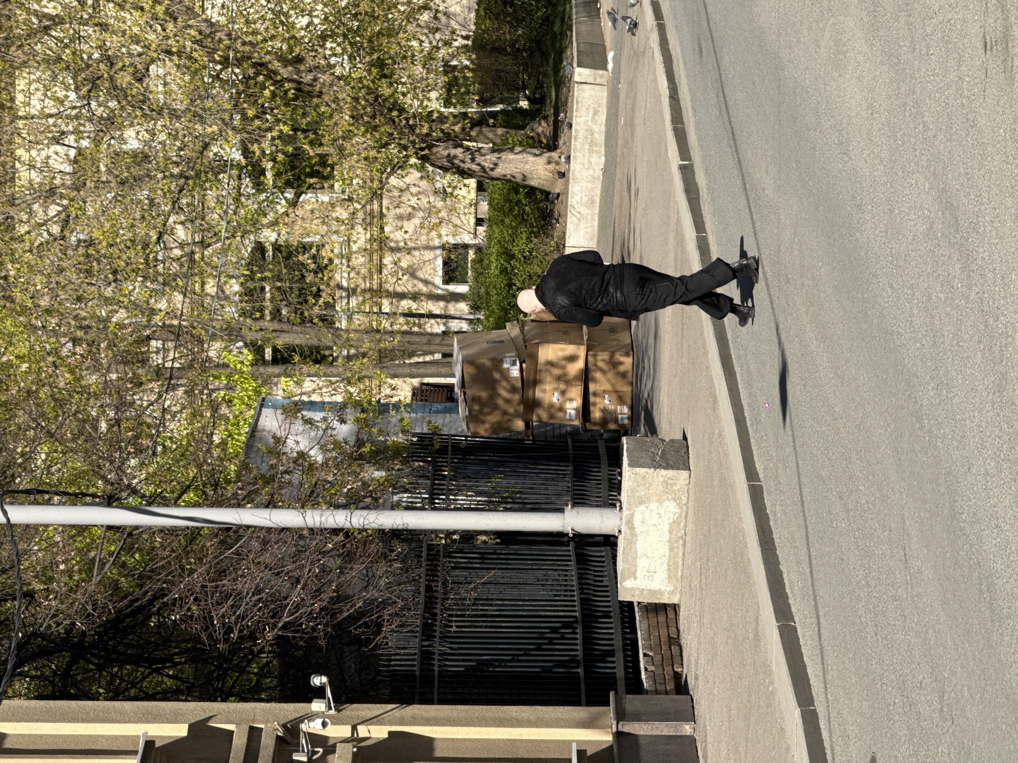 The image depicts a street scene in Moscow, Russia. A man in a black suit stands on a sidewalk next to a stack of cardboard boxes. He appears to be inspecting the boxes. A metal fence runs along the sidewalk, separating it from a tree-lined area and a building. The street is paved and appears to be a residential area. The time of day is daytime, with sunlight casting shadows. The environment consists of trees, buildings, a sidewalk, a street, and urban infrastructure such as security cameras on the side of a building.