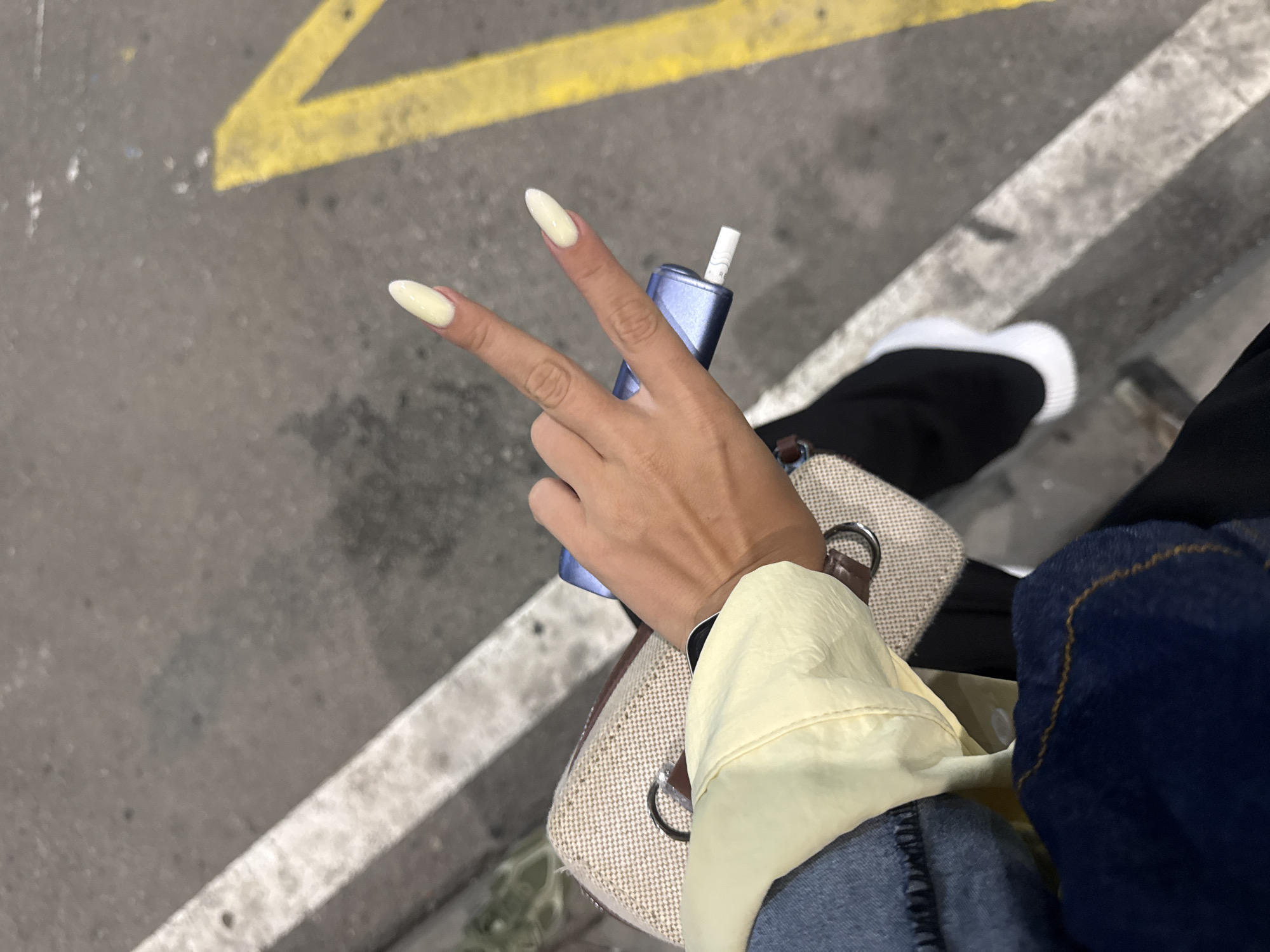 An overhead shot shows a person's hand holding a blue vape device and making a peace sign. The person is wearing light yellow nails. The background is a grey asphalt surface with a white line and a yellow arrow marking. A beige purse is visible in the lower left corner of the image. The person is wearing blue jeans and a light yellow jacket. The scene appears to be outdoors, possibly on a street or parking lot in Sofia, Bulgaria.