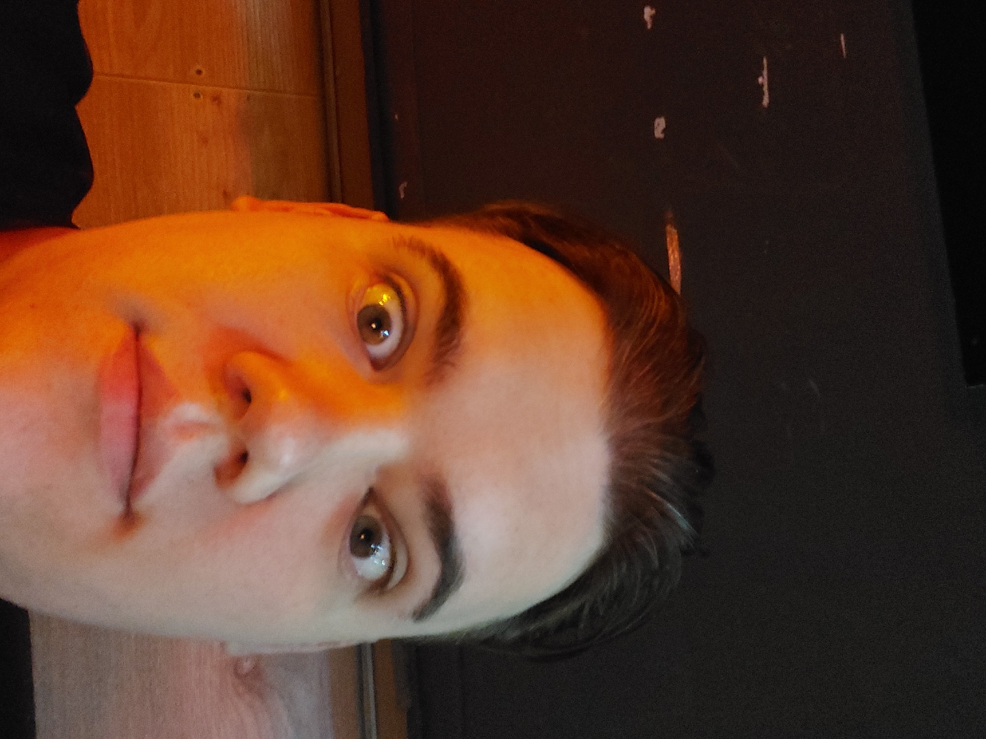 A close-up, slightly rotated image shows a person's face under warm, yellowish lighting. The person has fair skin, dark eyebrows, and light brown eyes with a glint of light reflecting in them. The person's hair is dark and neatly styled. The background is divided between a dark, possibly black wall with minor markings, and a lighter, yellowish-brown wooden surface. The location is undetermined, and there is no information available in the photo to indicate that it is in Tashkent, Uzbekistan.