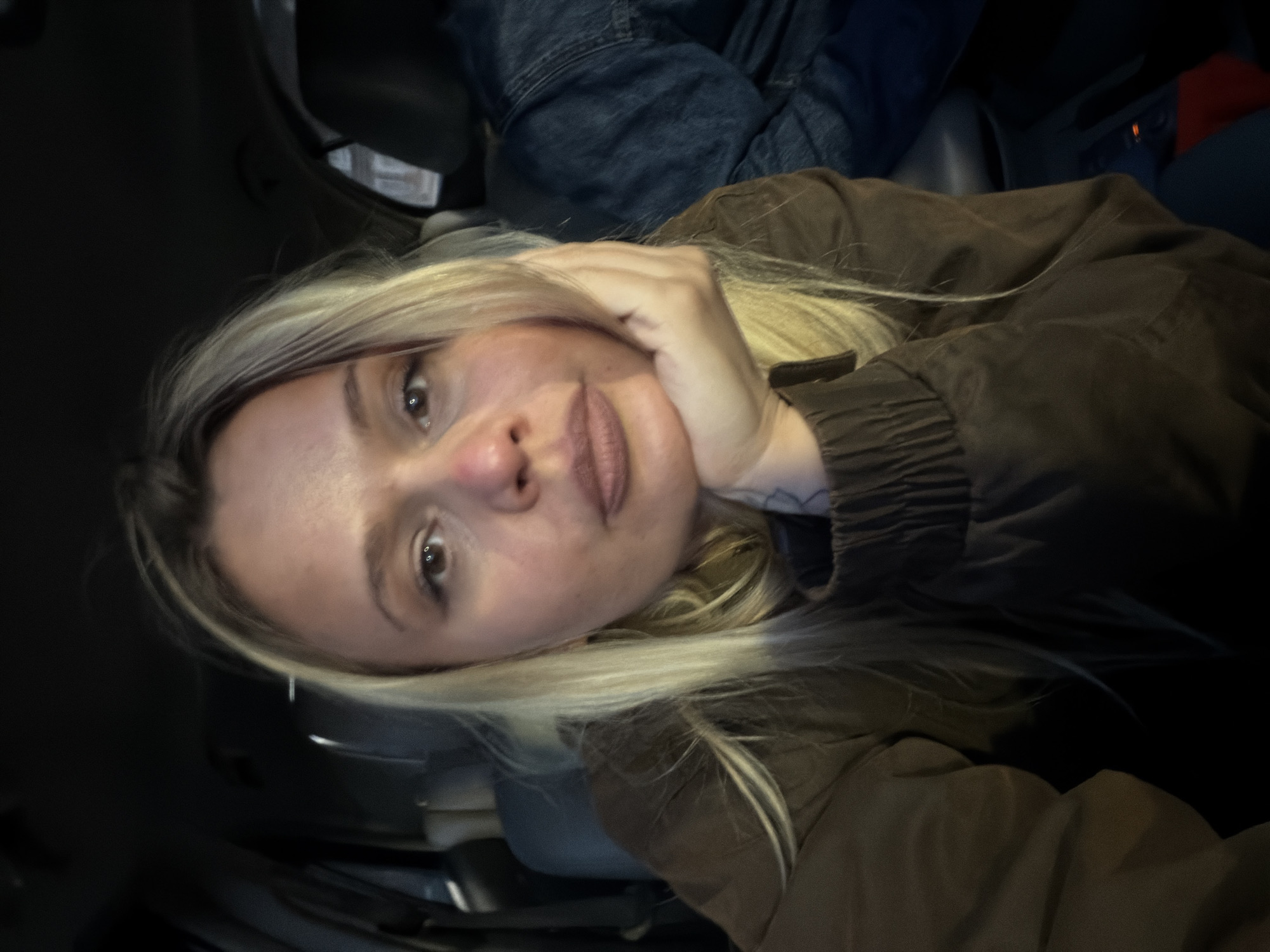 The image shows a woman with blonde hair resting her head on her hand. She appears to be inside a vehicle. There is no visible indication of the location being in Sarapul, Russia. The environment is dimly lit.