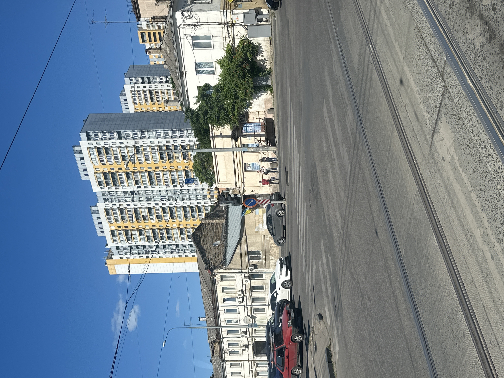 This media file shows a street scene in Odesa, Ukraine. The image is taken from a slightly elevated perspective, looking down a street with tram tracks. On the left side of the street, there are several tall apartment buildings with a mix of yellow and white facades. These buildings are modern and appear to be residential.
In contrast, closer to the foreground on the right side of the street, there are older, lower-rise buildings. These appear to be made of concrete and plaster and show signs of age. Between these different styles of architecture, a pedestrian crossing is visible on the road.
Several cars are parked along the street, including a red hatchback, a black car, a white sedan, and a dark grey car further down the road. A few people are standing on the sidewalk near the older buildings, suggesting the scene is active, though not crowded.
The sky is a clear, bright blue, indicating a sunny day, likely midday given the strong shadows cast by the buildings and the bright light. There are some power lines and a traffic signal visible. No specific events are clearly depicted, but the general atmosphere is that of a typical urban environment. The tram tracks suggest this is a route for public transportation.