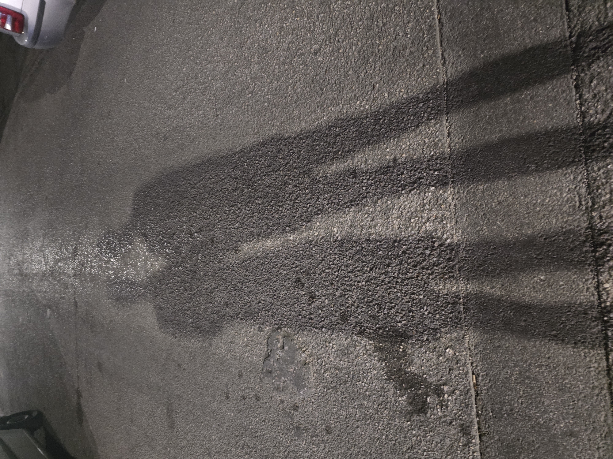 The image shows two shadows cast on wet asphalt at night. The shadows appear to be those of two people standing close together. The left shadow is smaller and seems to be of a person with their arm around the taller shadow's shoulder. The shadows are elongated due to the low angle of the light source. The asphalt is dark and reflective in places, indicating recent rain or dampness. In the background, the rear bumper and taillight of a silver car are visible on the right side of the image. The overall impression is a moment captured in a city street in Nea Ionia, Greece, under artificial lighting.